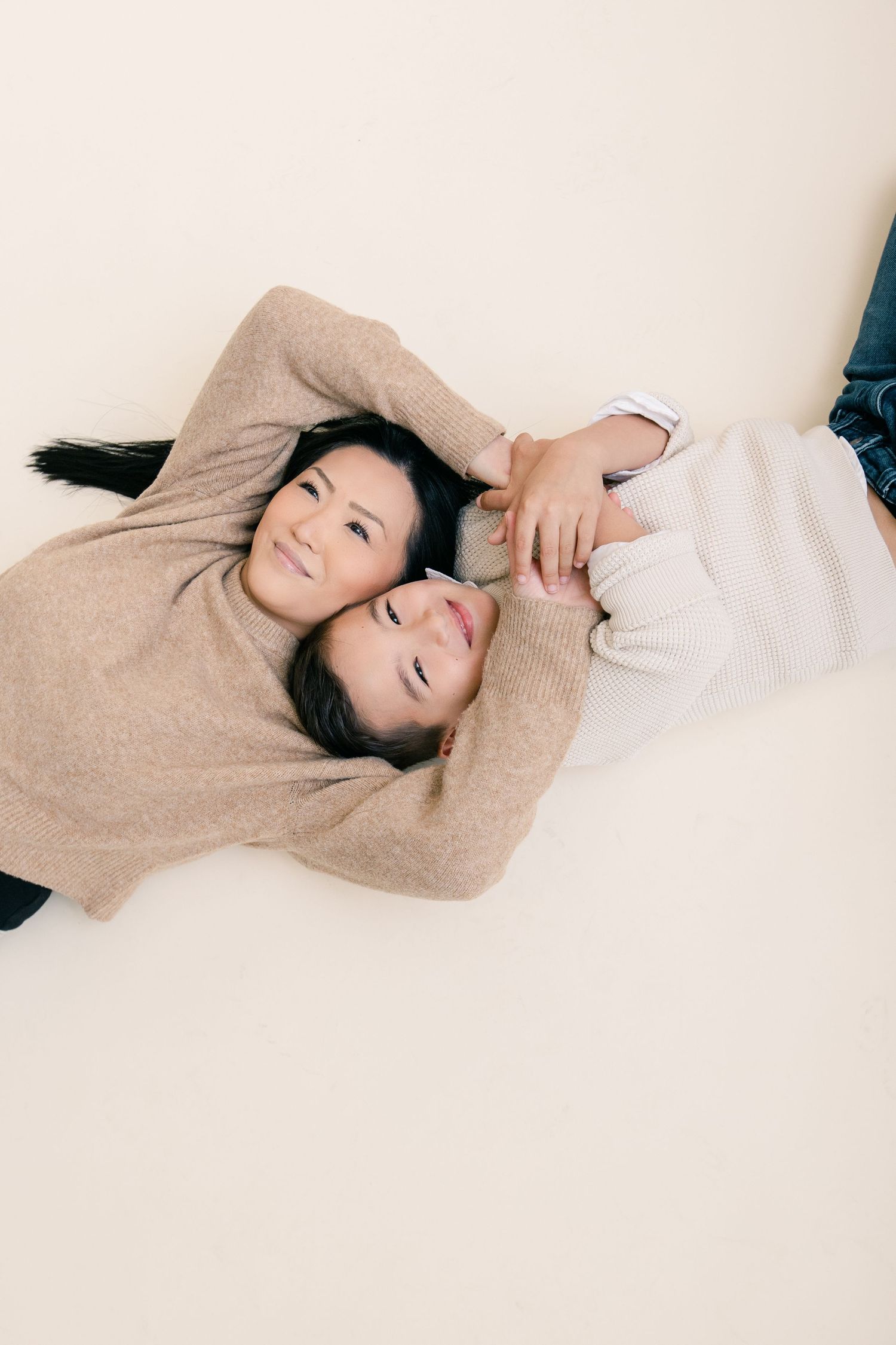Two women in cozy sweaters lying on a light background, smiling and embracing in a warm, candid portrait.