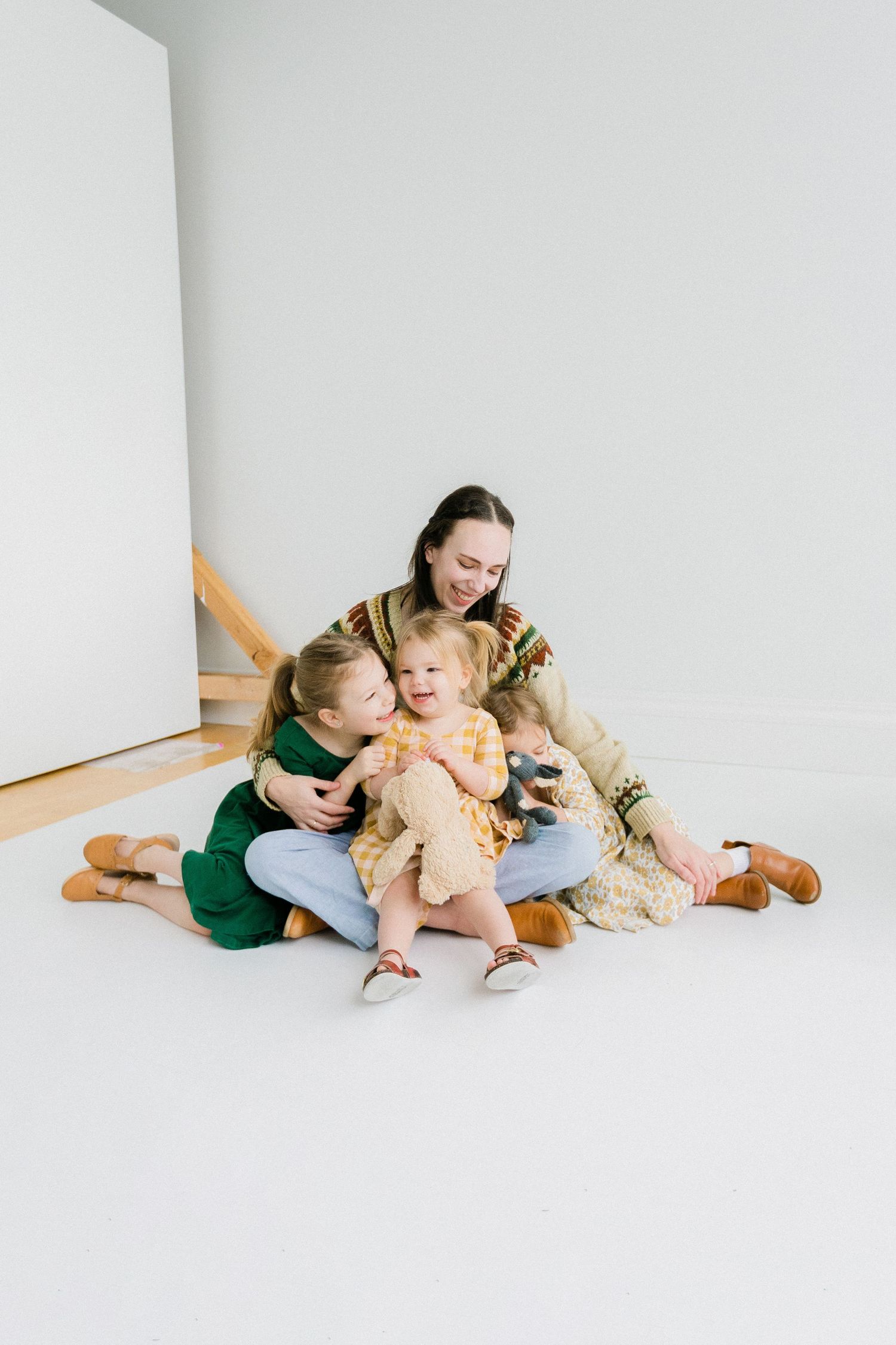 A mother sits on a white floor with two young daughters and a small dog, sharing a warm family embrace.