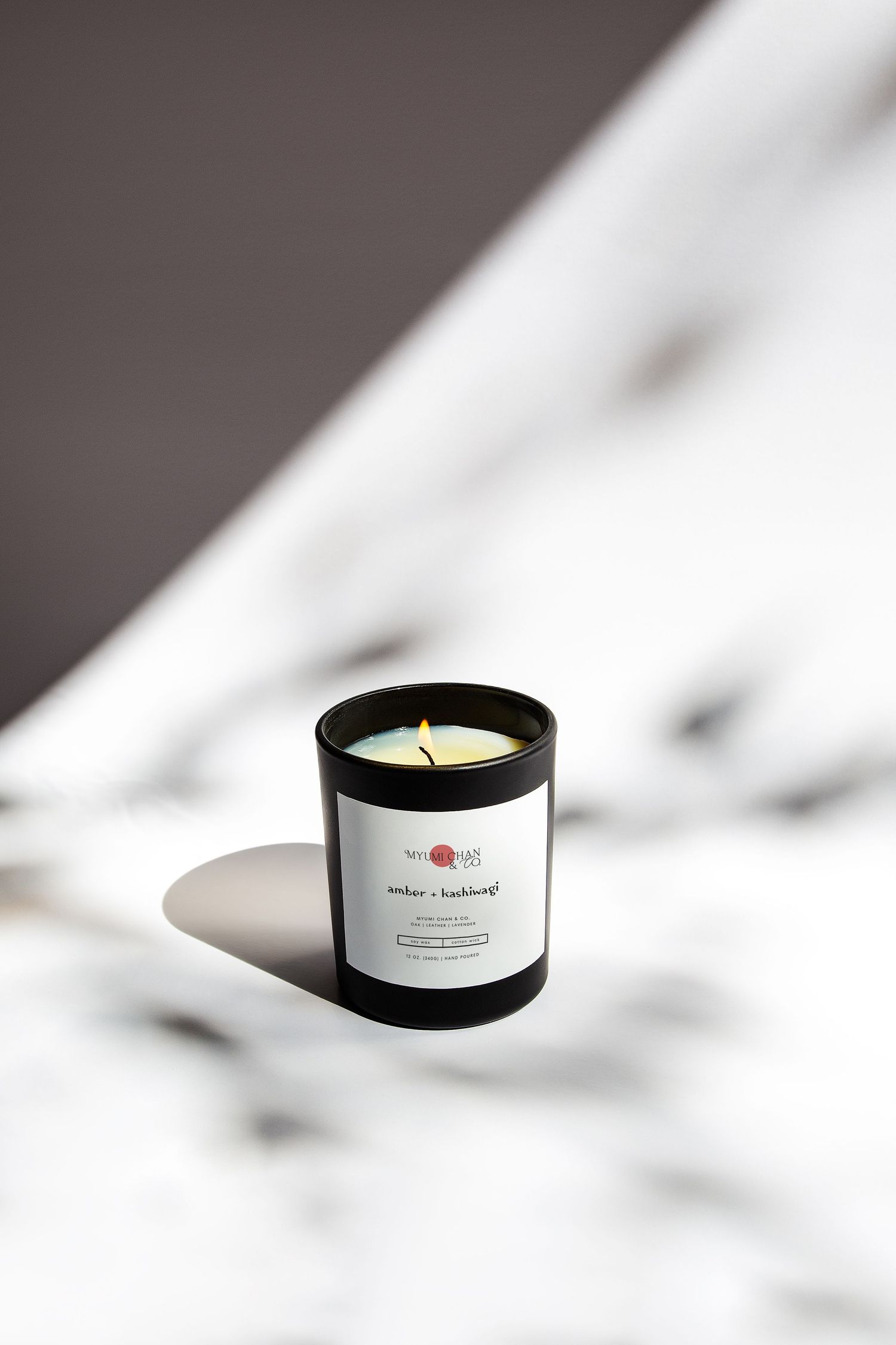 Black glass amber and patchouli scented candle with white label, lit wick, on marble surface with dramatic shadows.