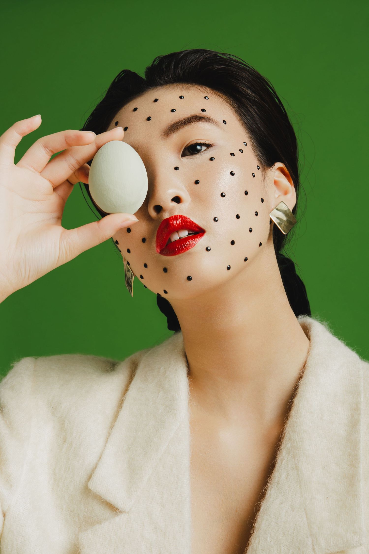 Asian woman with red lips, black dot face art, holding a green egg, wearing white blazer on green background.