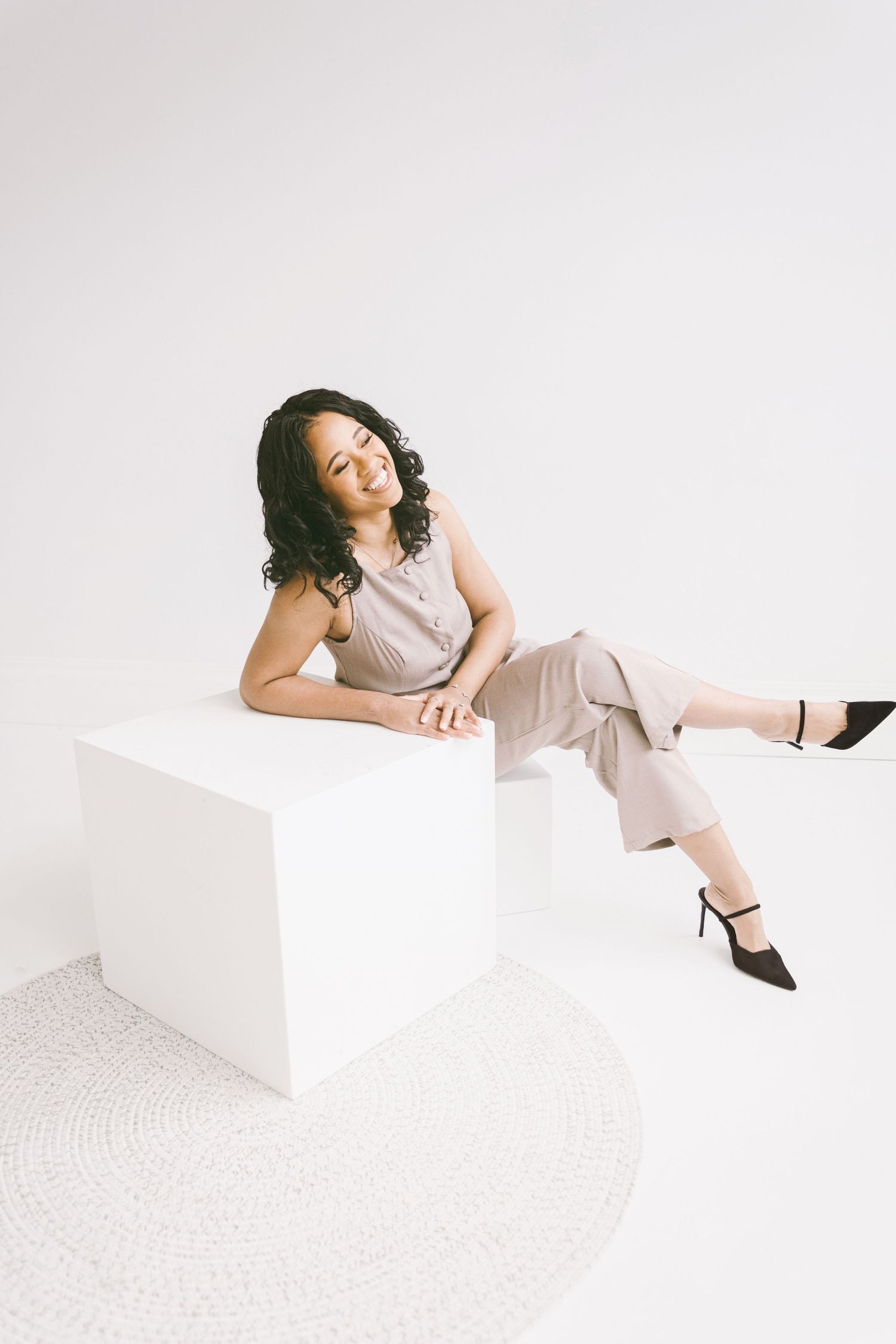A young woman in a beige jumpsuit and black heels sits relaxed on a white cube against a white background.