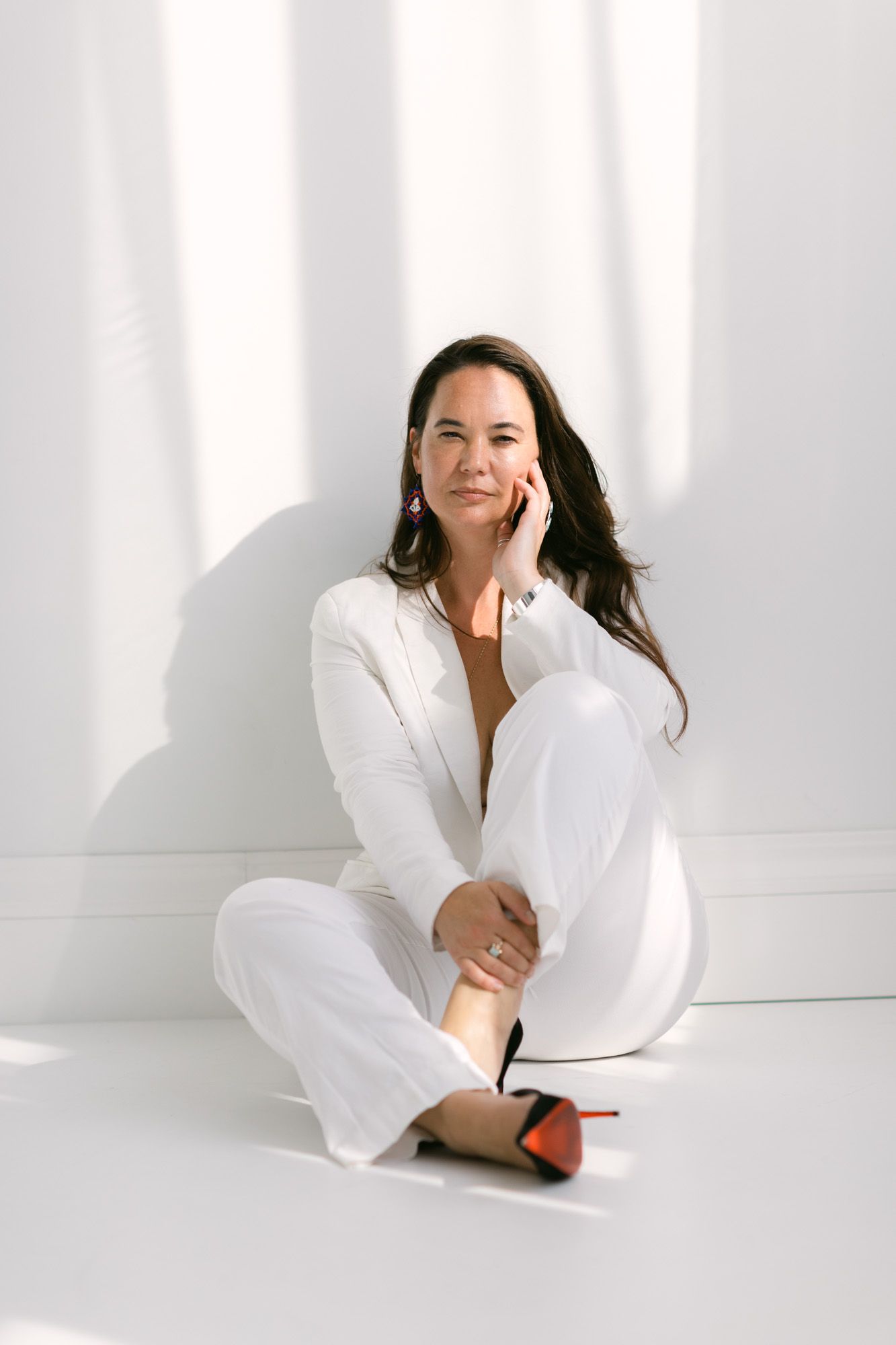 Woman in white suit sitting cross-legged on floor, resting hand on cheek, wearing red-soled heels.