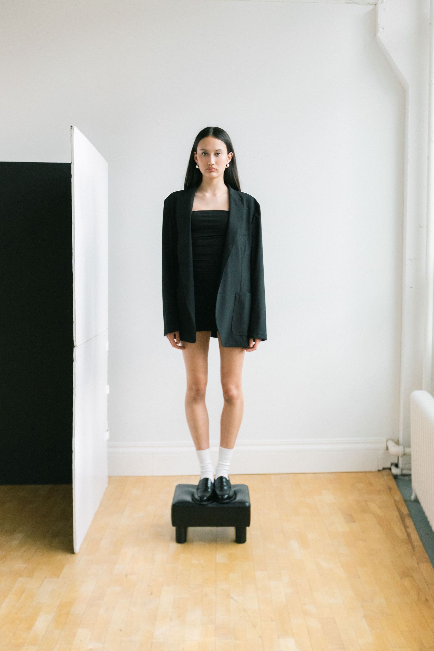 Woman in oversized black blazer and mini dress with white socks and loafers standing on a platform.