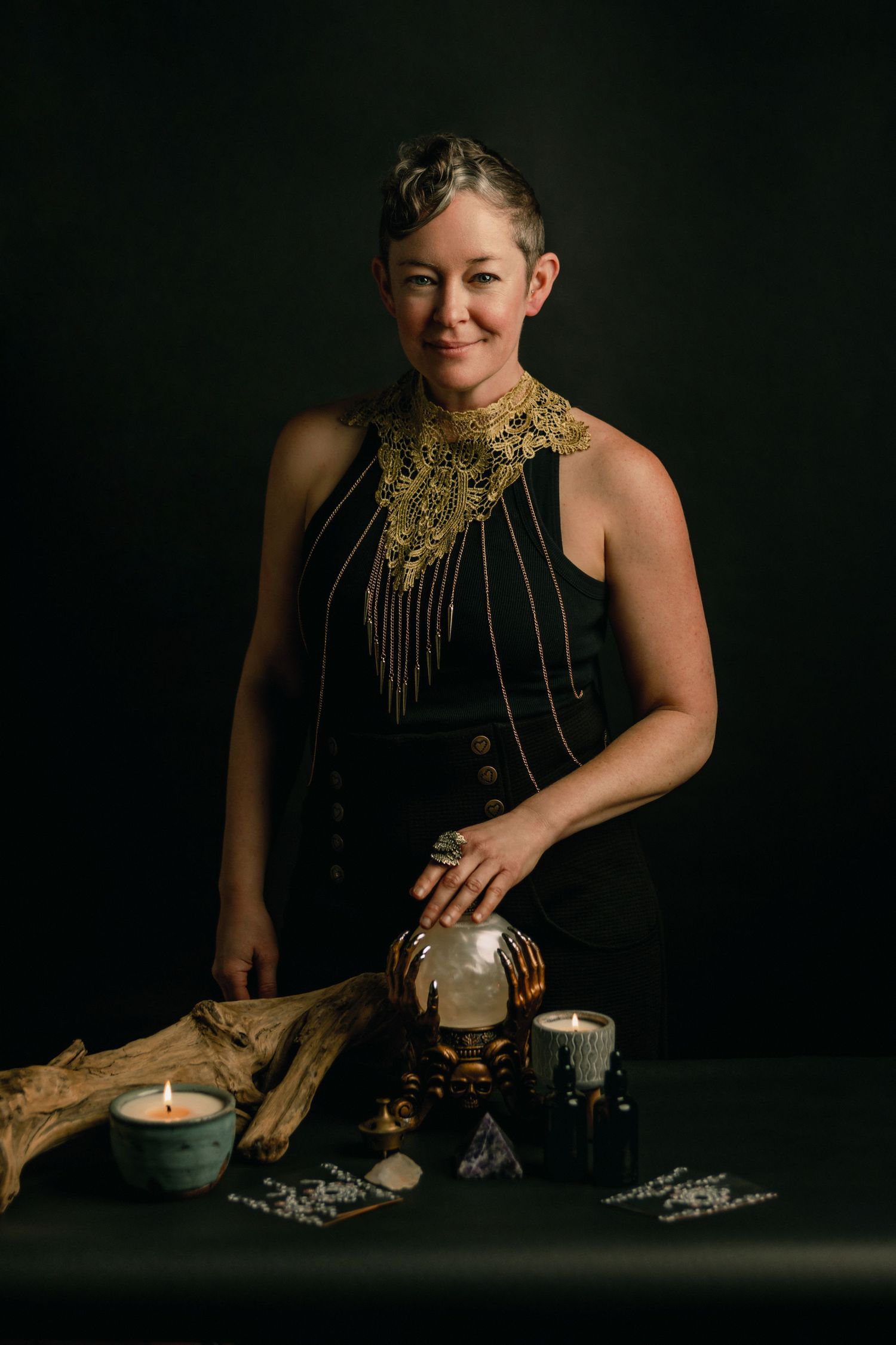 Woman in black halter top with gold lace collar poses with crystal ball, candles, and driftwood on dark background.