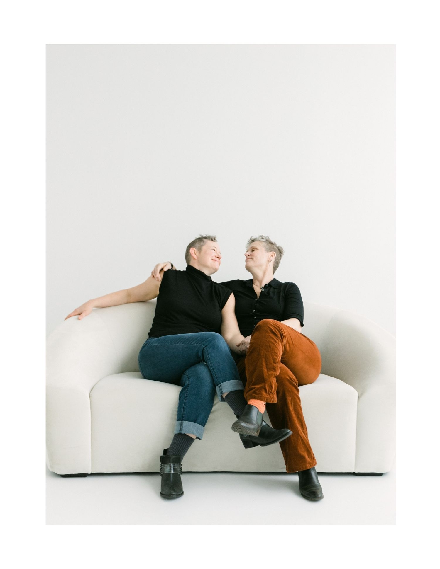 Two women sitting close on a white sofa, laughing and gazing at each other in a bright studio.