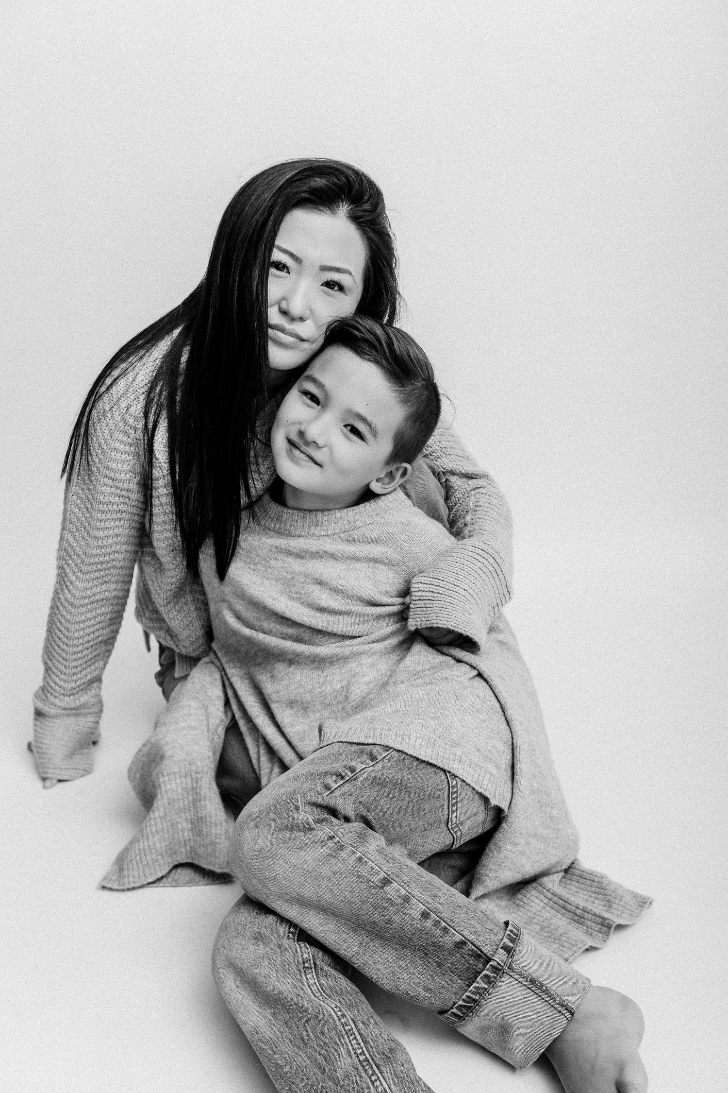 Black and white photo of a smiling woman hugging a young boy, both wearing cozy sweaters, sitting together on a white background.