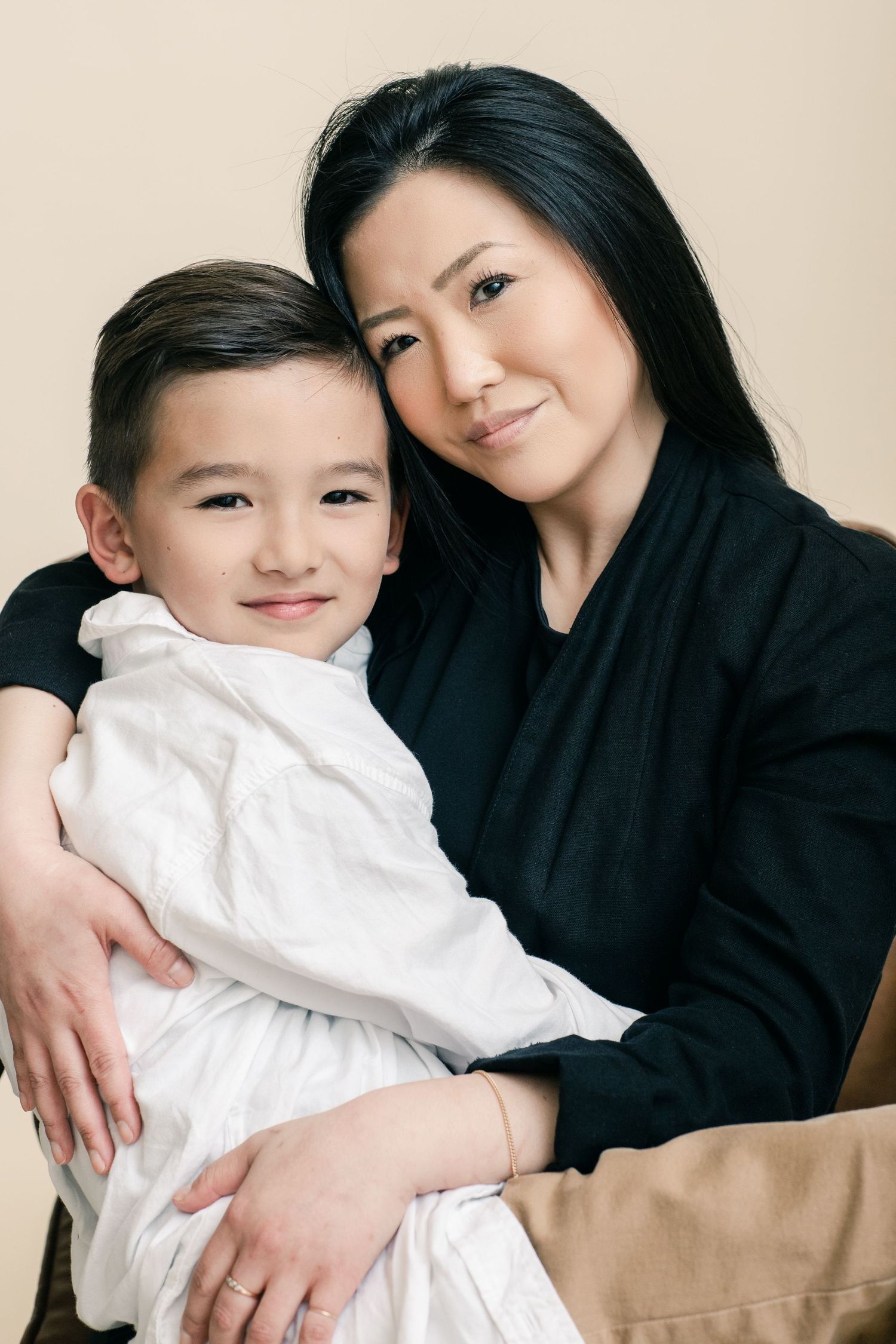 A woman with long black hair hugs a young boy in white, both smiling warmly in a studio portrait.