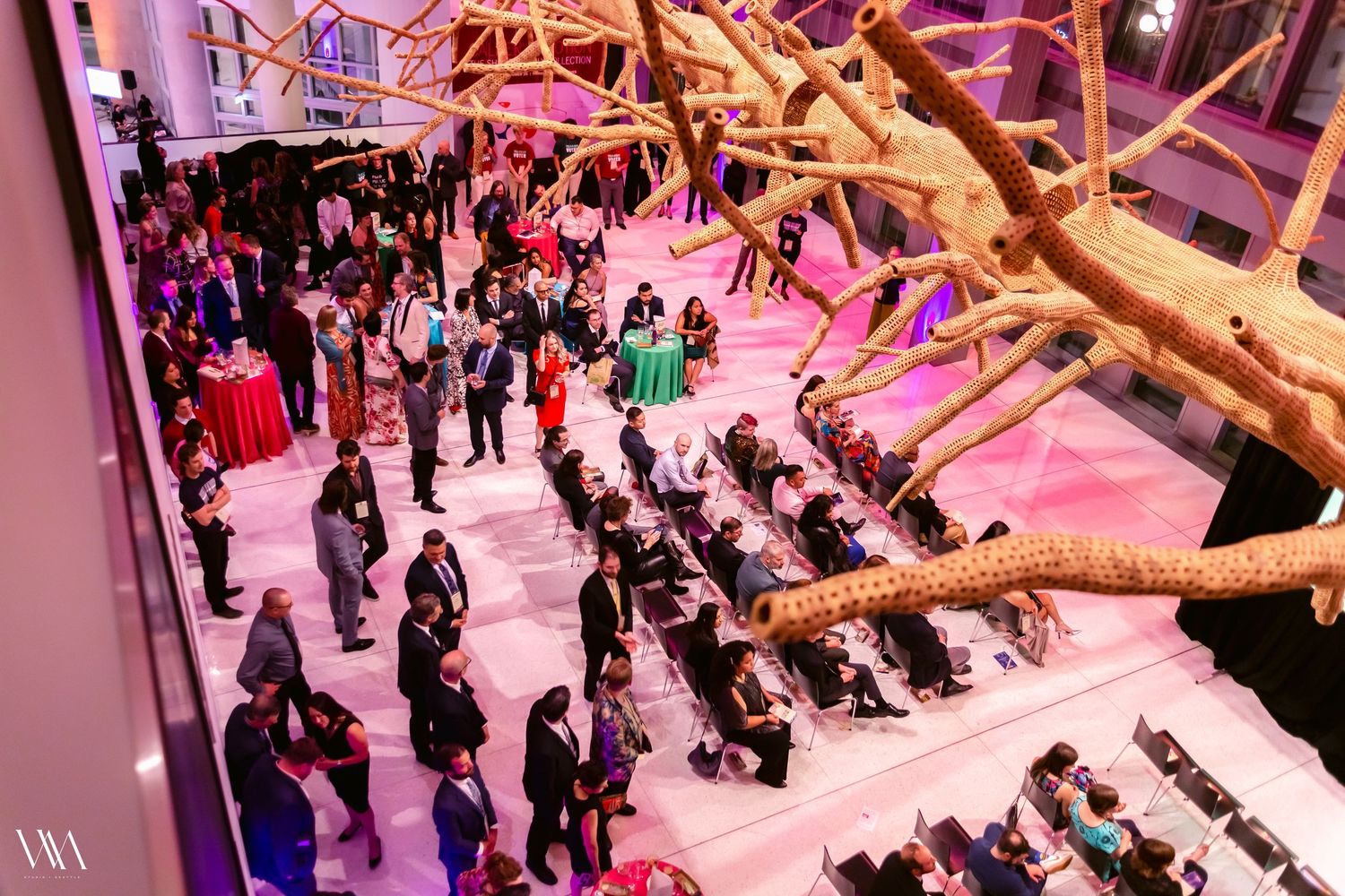 Aerial view of an elegant indoor event with guests mingling under a decorative tree installation with pink lighting.