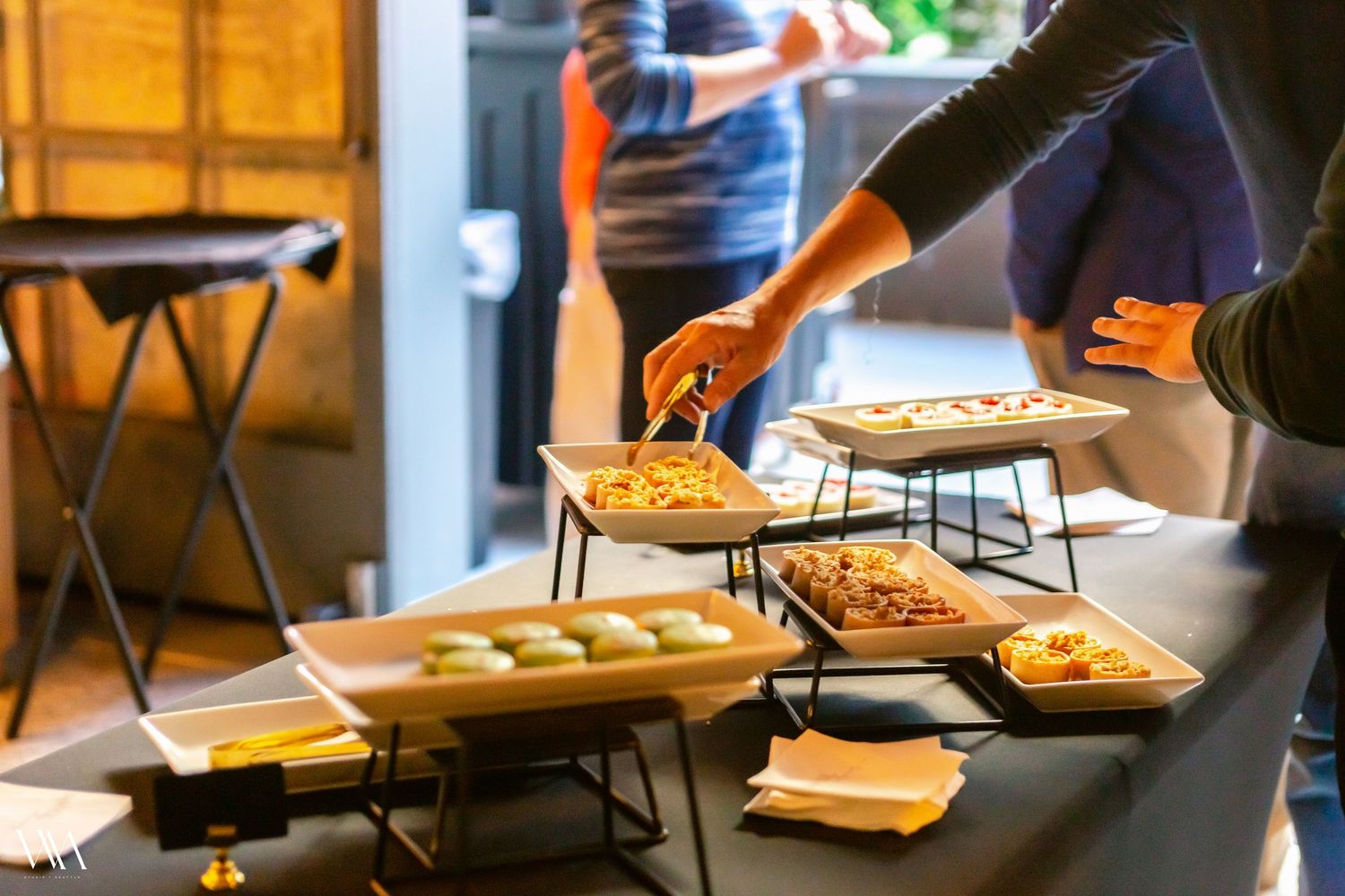 People serving food at a buffet table with multiple dishes in warming trays at a catered event.