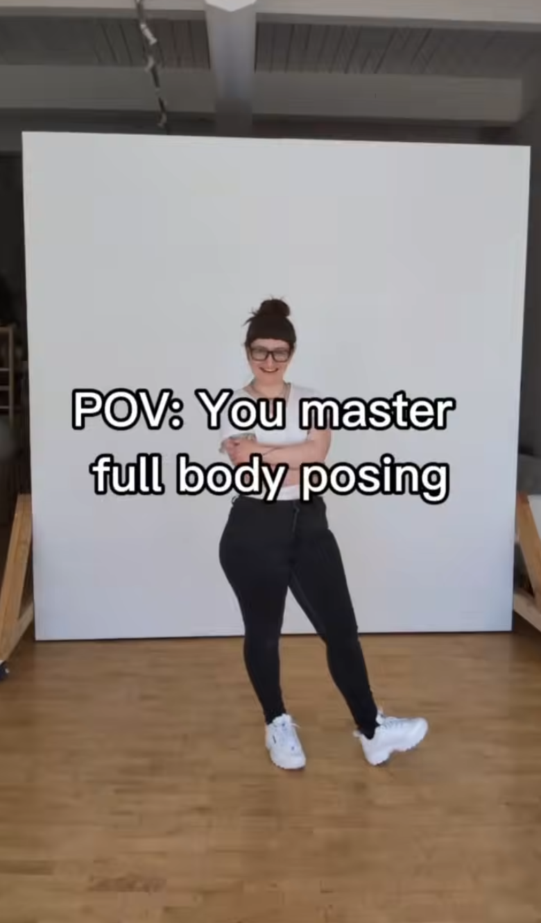 Woman in black outfit demonstrates full body posing techniques in a studio with white backdrop and wooden floor.