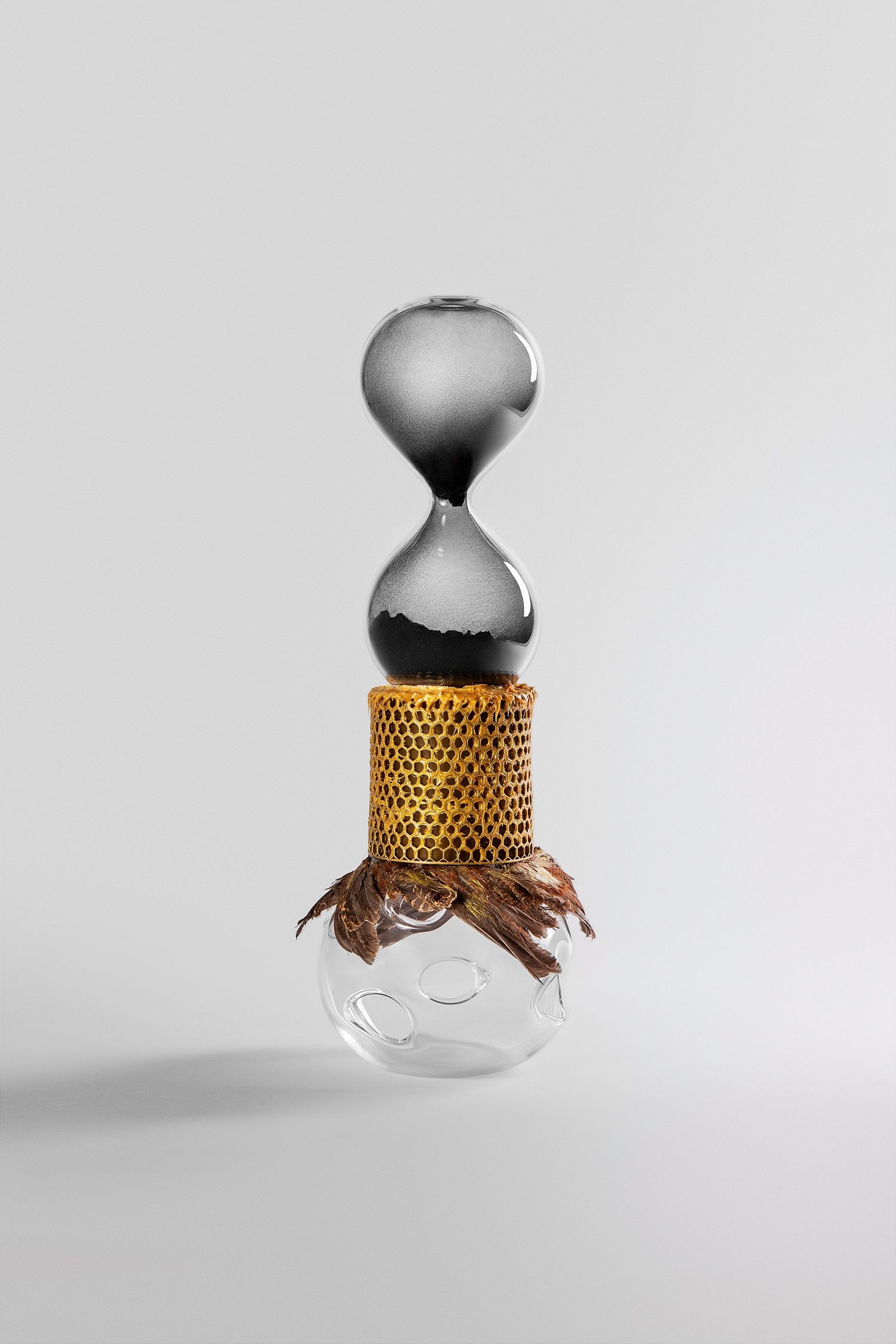 Surreal sculpture combining a dark hourglass, golden honeycomb cylinder, and clear glass bulb with dried grass on white background.