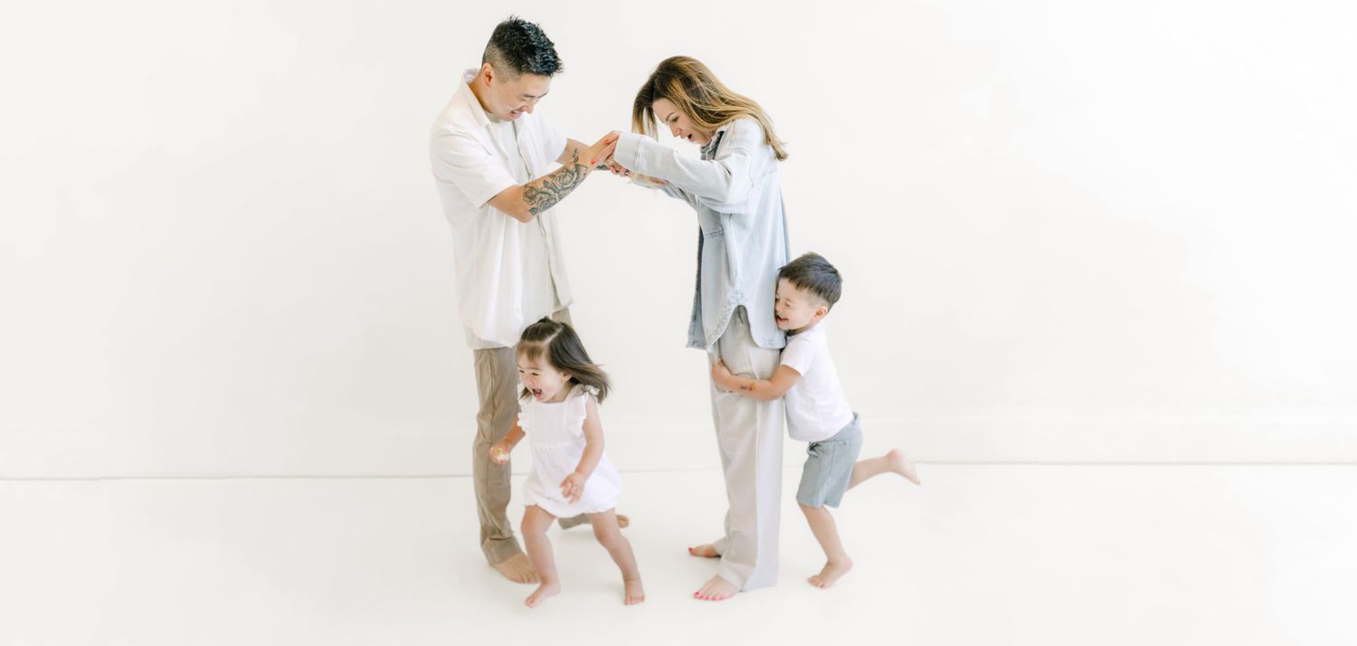 Asian family of four dancing and playing together against a white background, parents holding hands with toddler children.