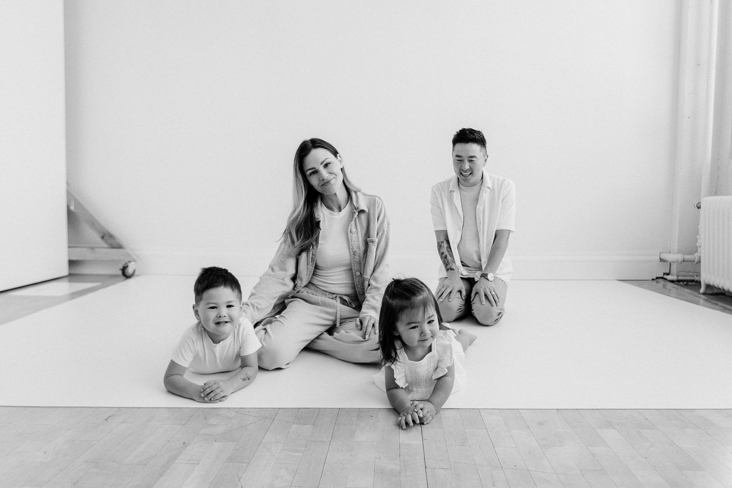 Black and white family photo of a woman, man, and two toddlers sitting on a white studio floor.
