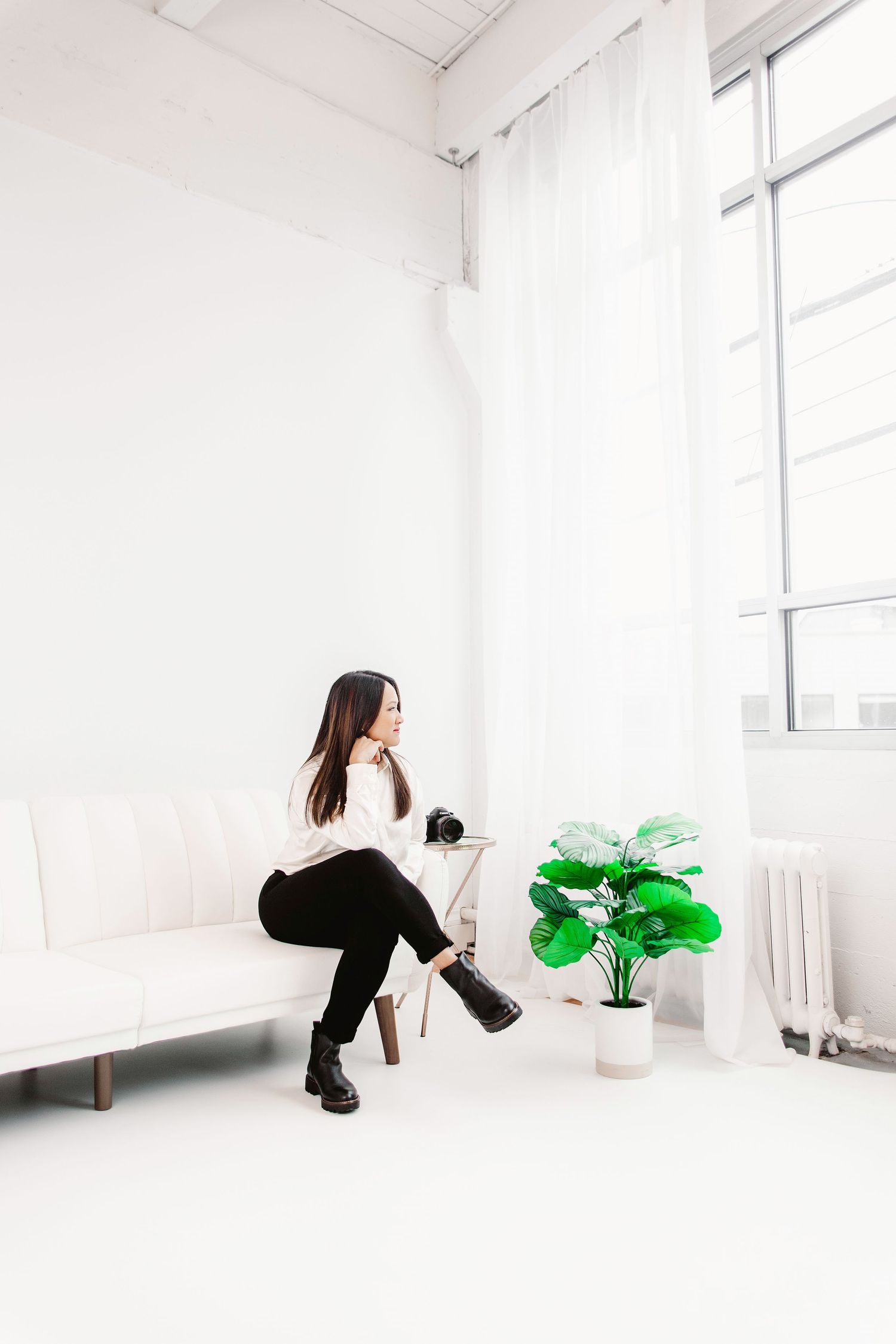 Woman in white top and black pants sits on a white sofa holding a camera near a green plant by large windows.