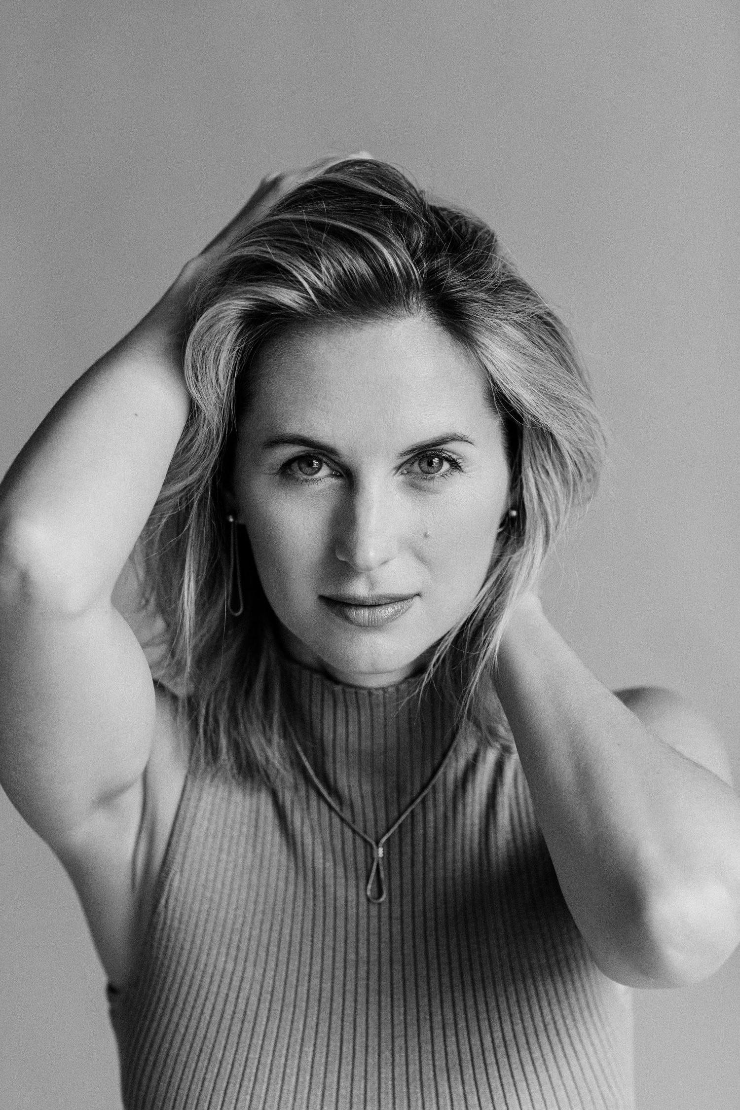 Black and white portrait of a blonde woman in a ribbed top, posing with hands in hair, wearing a delicate necklace.