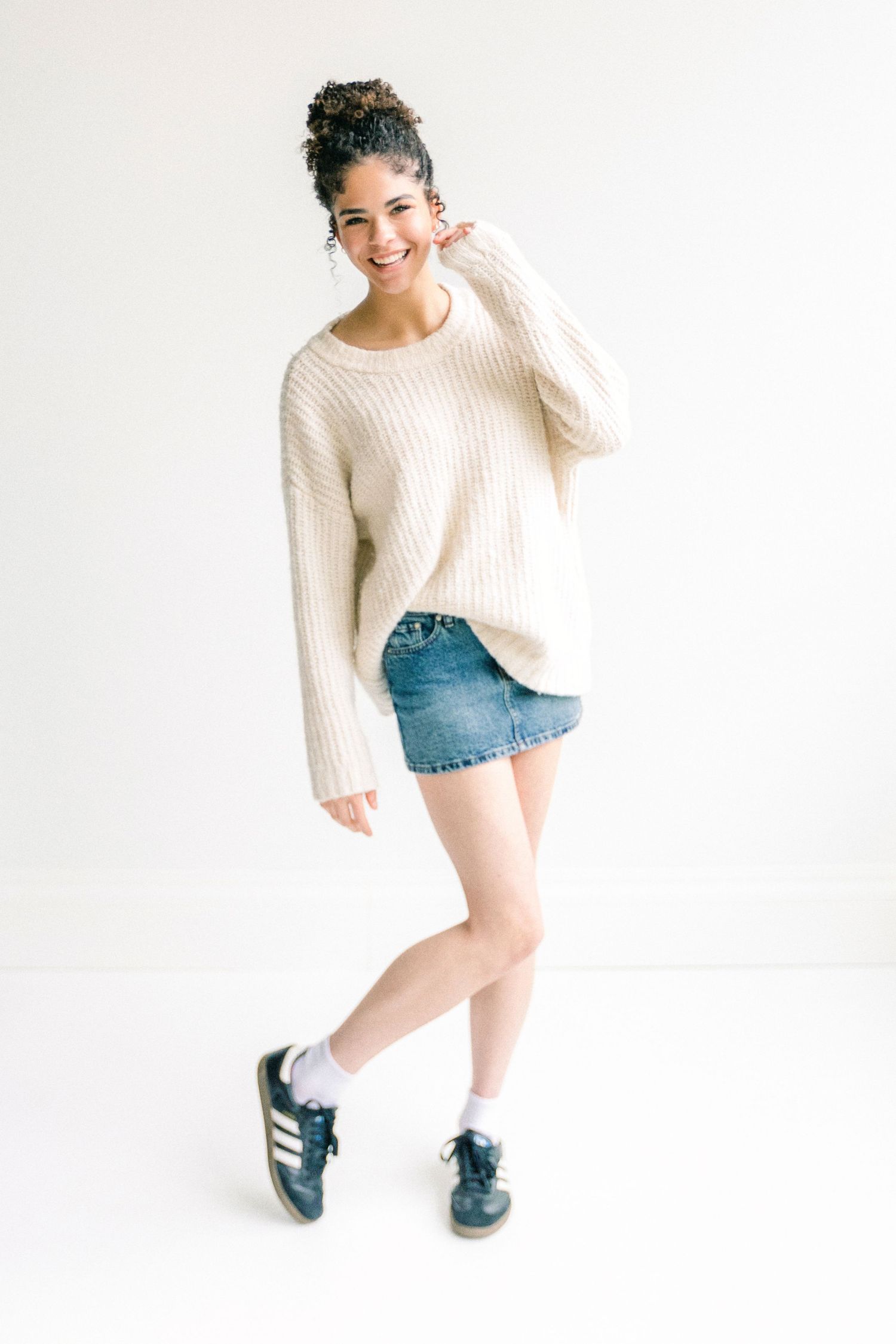 Young woman smiling in cream knit sweater, denim mini skirt, and Adidas sneakers posing against white background.