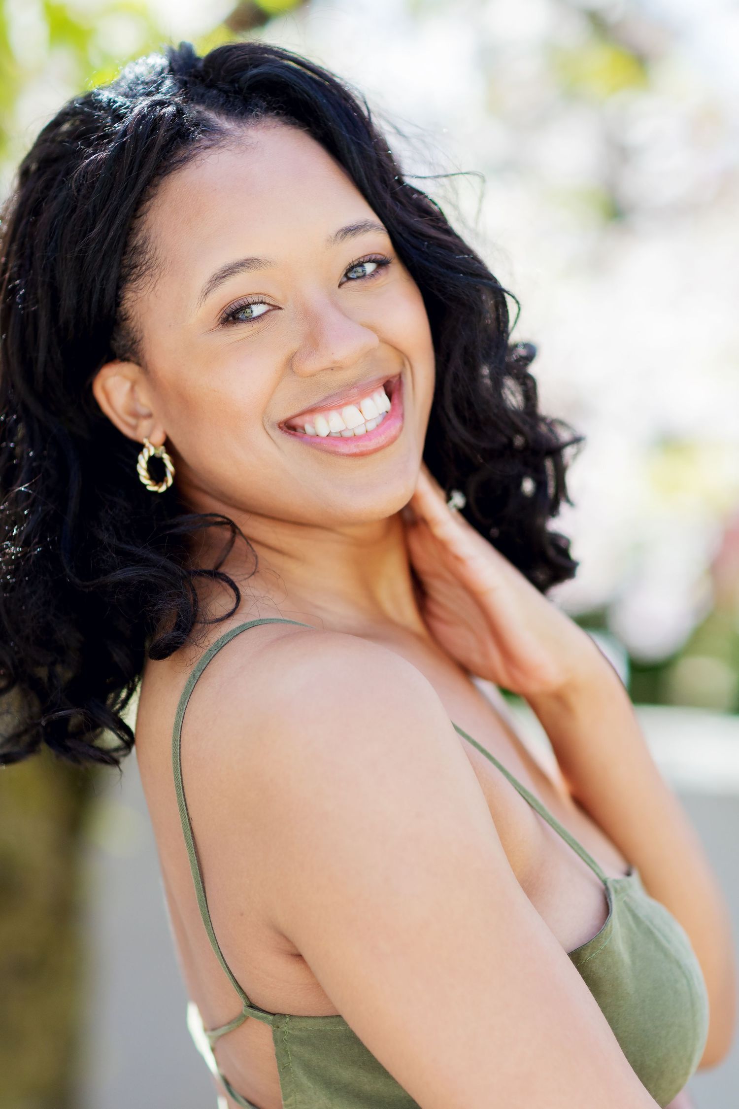 Smiling young woman with curly hair wearing gold earrings and green top outdoors.