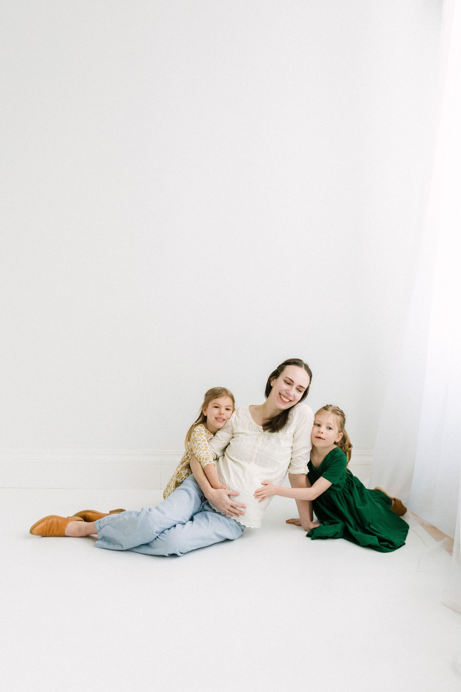 Pregnant woman in white dress sits on floor with two young girls in floral and green dresses in bright white room.