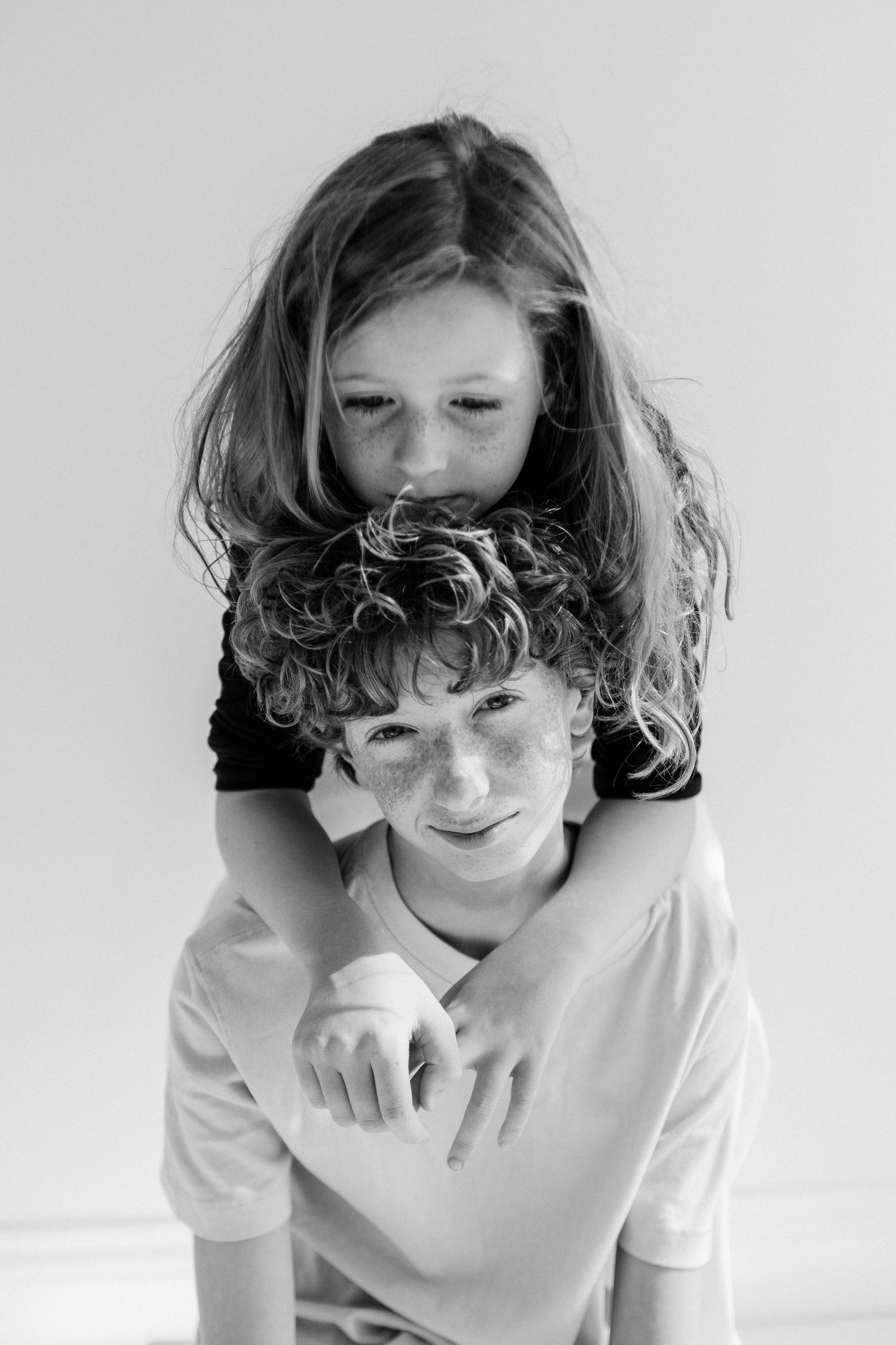 A black and white portrait of a curly-haired freckled boy with a girl leaning on his shoulders.