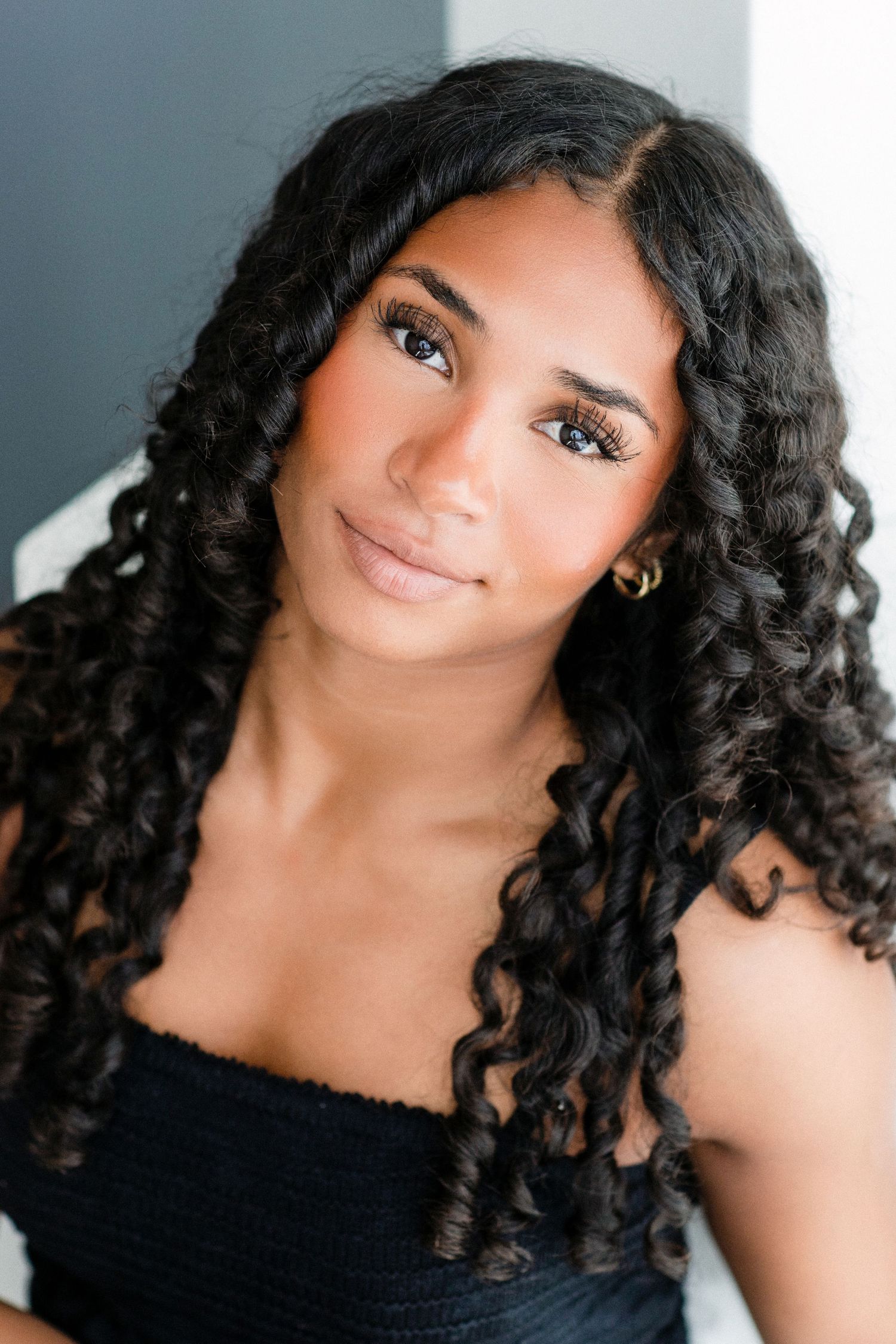 Young woman with long curly black hair, blue eyes, and gold hoop earrings wearing a black top.