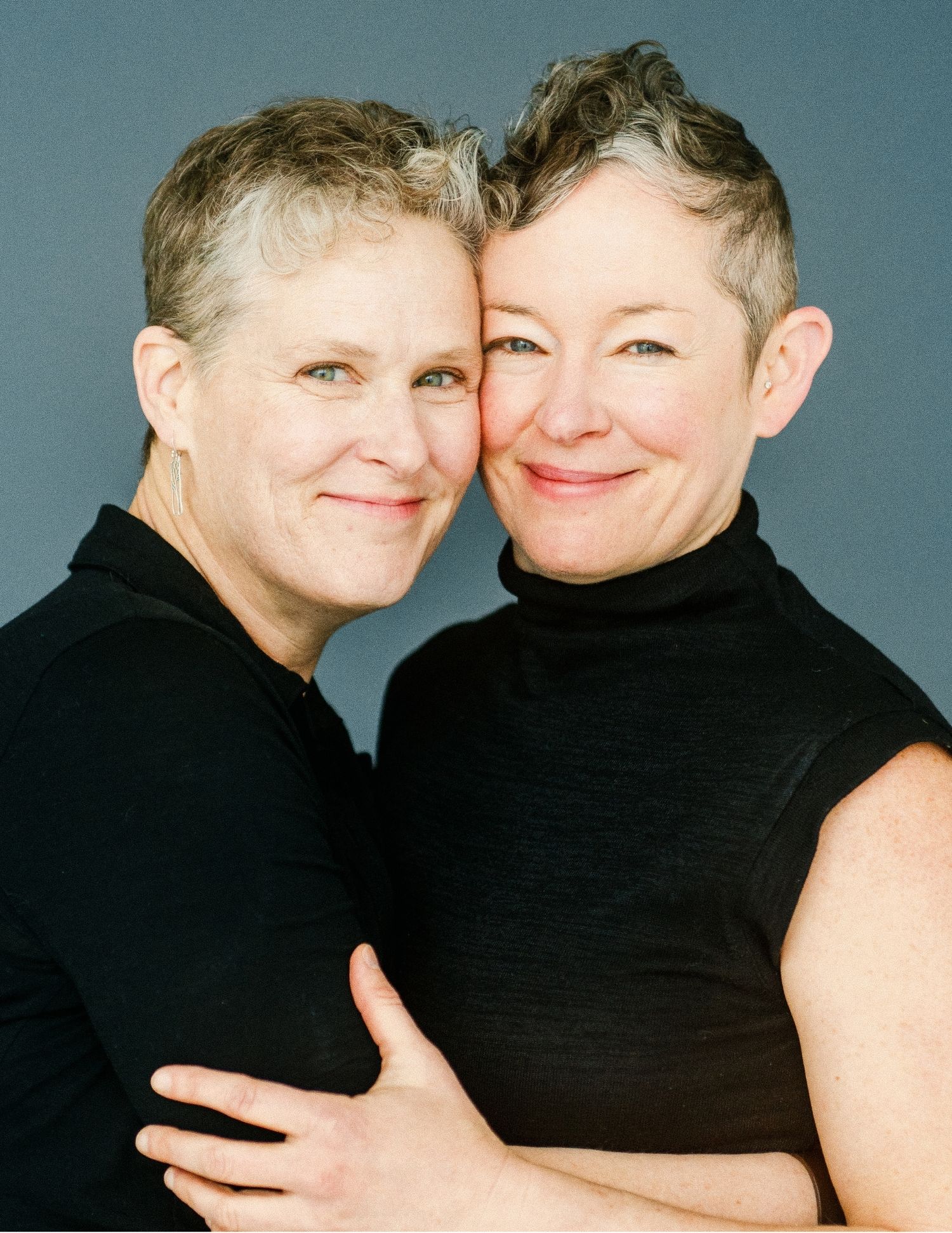 Two women with short gray hair embrace warmly, both wearing black tops, smiling in a studio portrait.