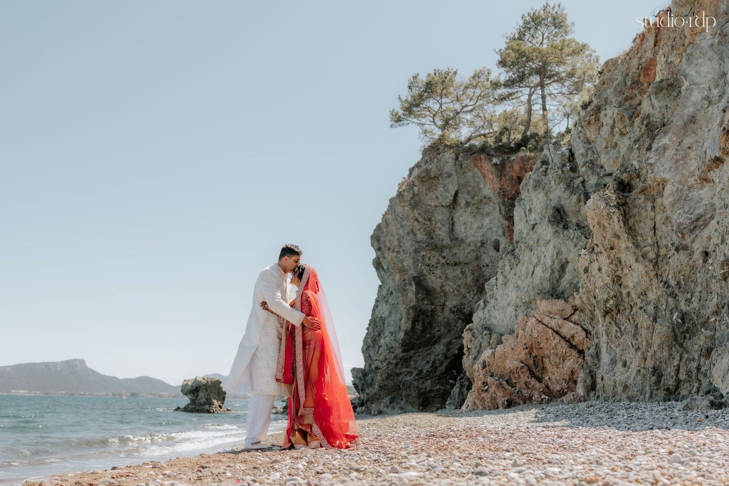 Couple in traditional Indian wedding attire sharing a romantic moment on a rocky Mediterranean beach shore.