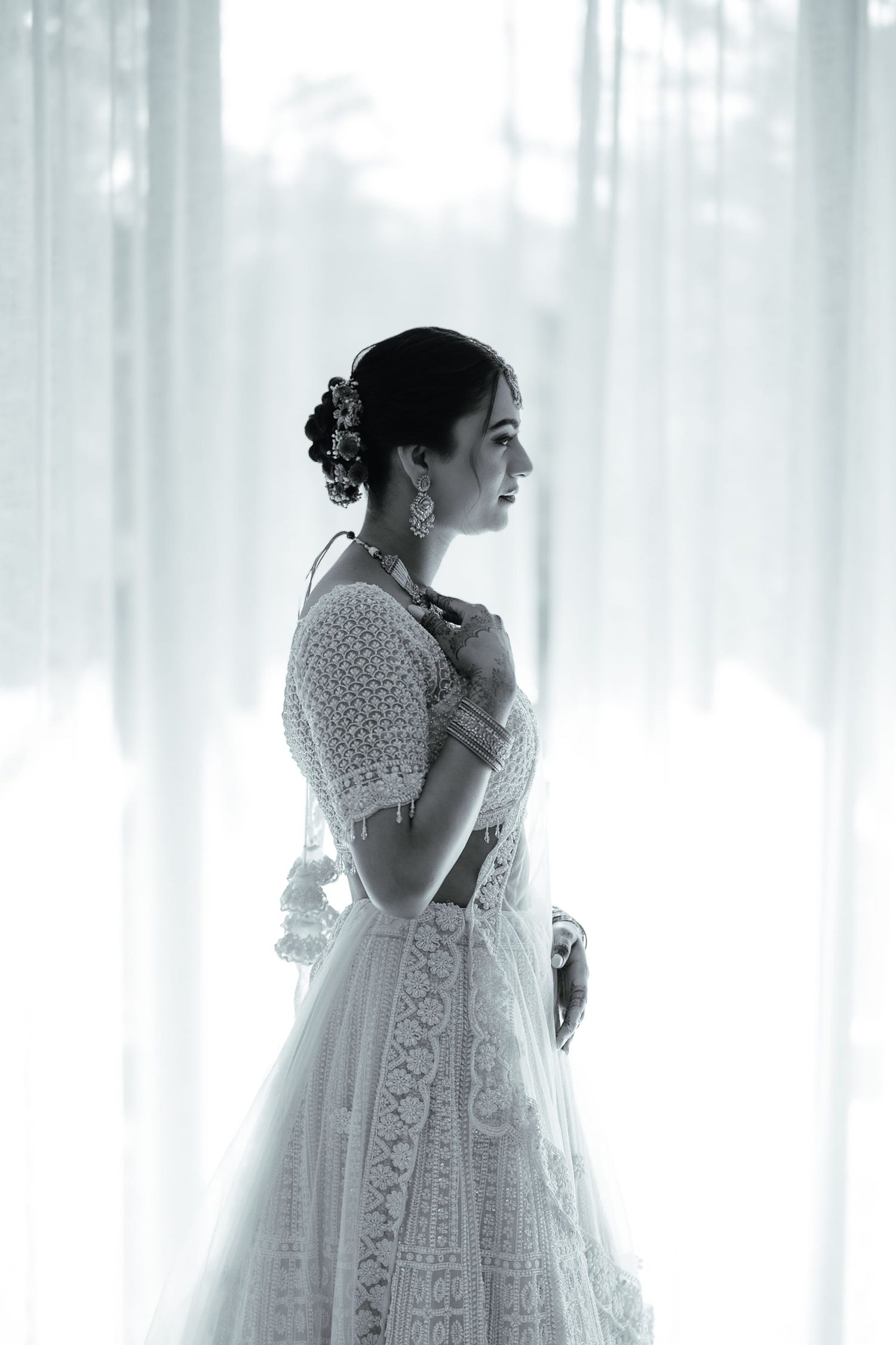 Black and white portrait of a bride in white lace dress with floral hair accessories standing by sheer curtains.