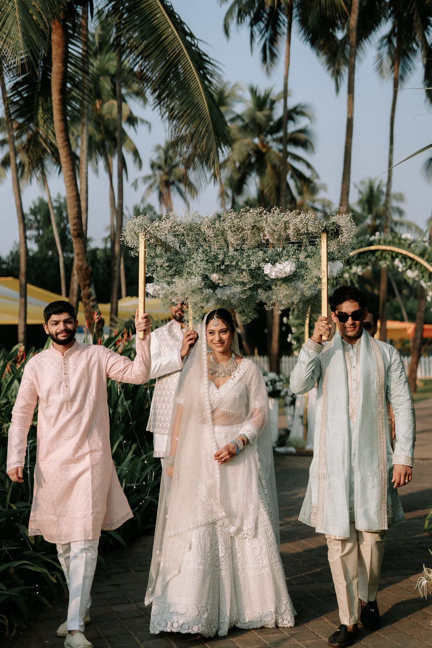 Bride in white lehenga walks the aisle flanked by two groomsmen in pastel sherwanis under a floral canopy.