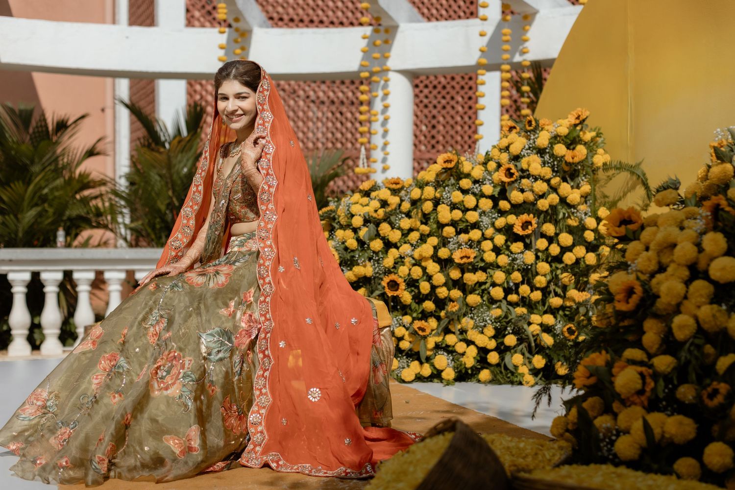 Bride in peach-colored lehenga poses elegantly among yellow flowers and white pillars during wedding photoshoot.