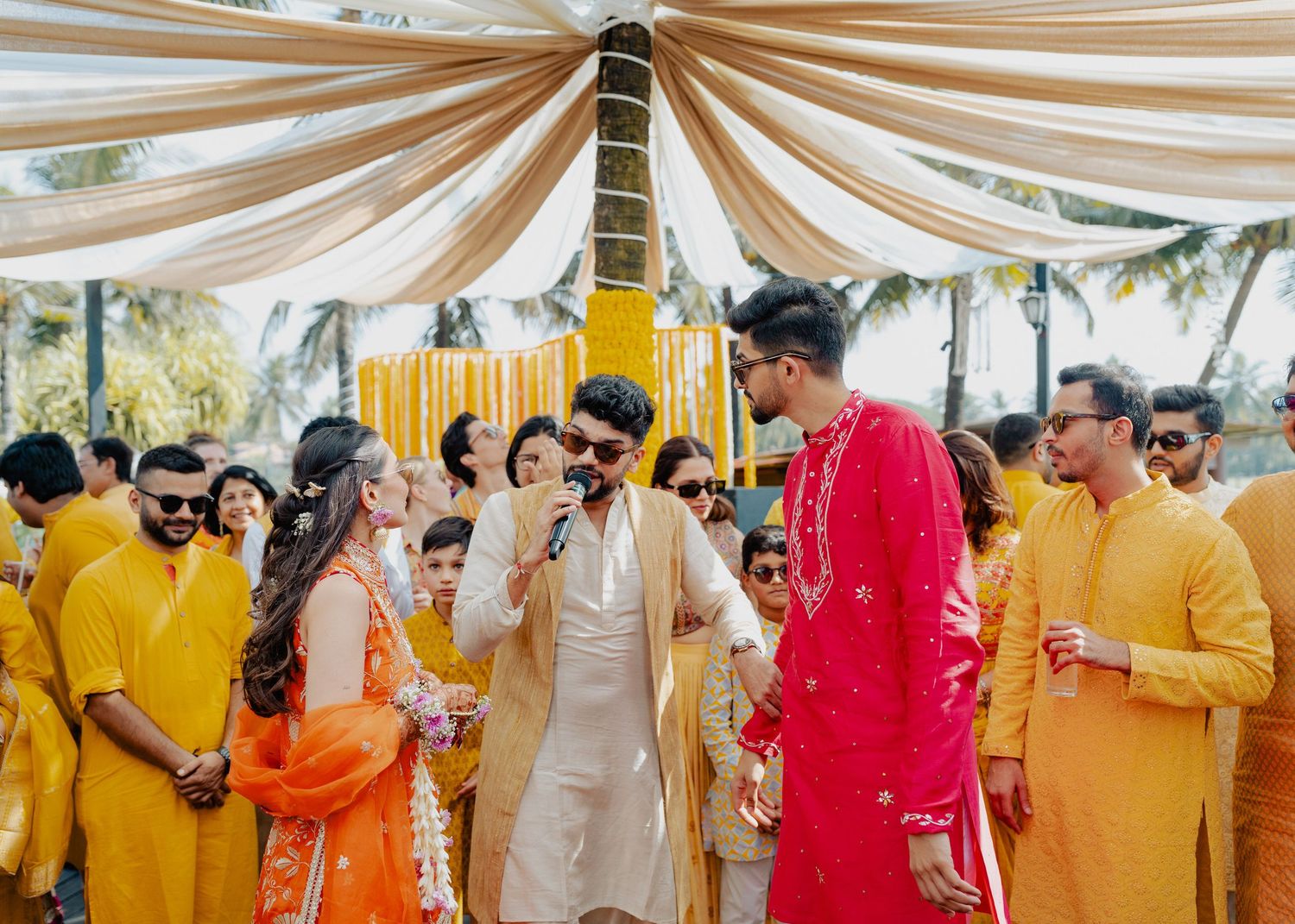 Traditional Indian wedding celebration with guests in vibrant yellow, orange and red outfits under draped white fabric canopy.