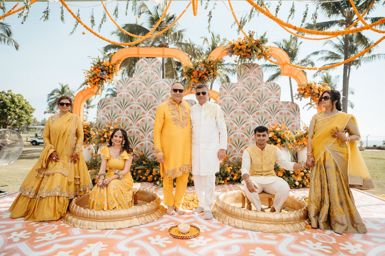 Traditional Indian wedding party in yellow and gold outfits posing at an outdoor ceremony with floral decorations.
