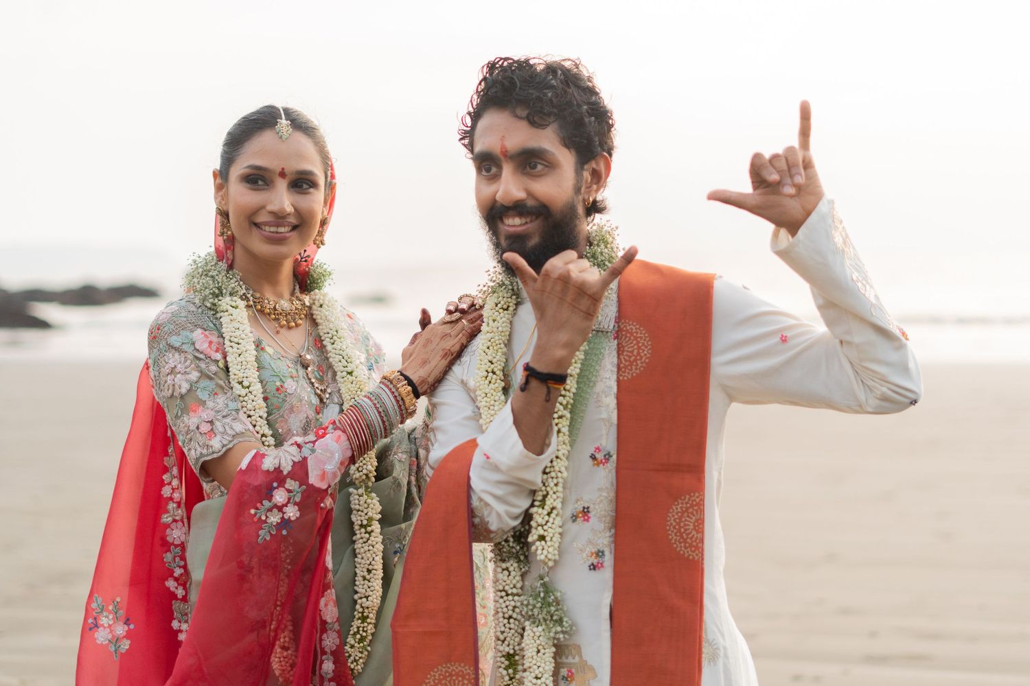 Indian couple in traditional wedding attire smiling on beach, bride in red lehenga with floral garland, groom in white kurta.