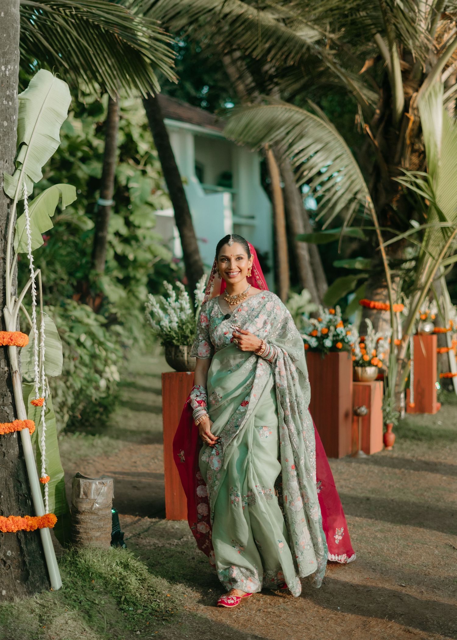 Bride in mint green saree with pink dupatta walks through tropical garden decorated with marigolds.