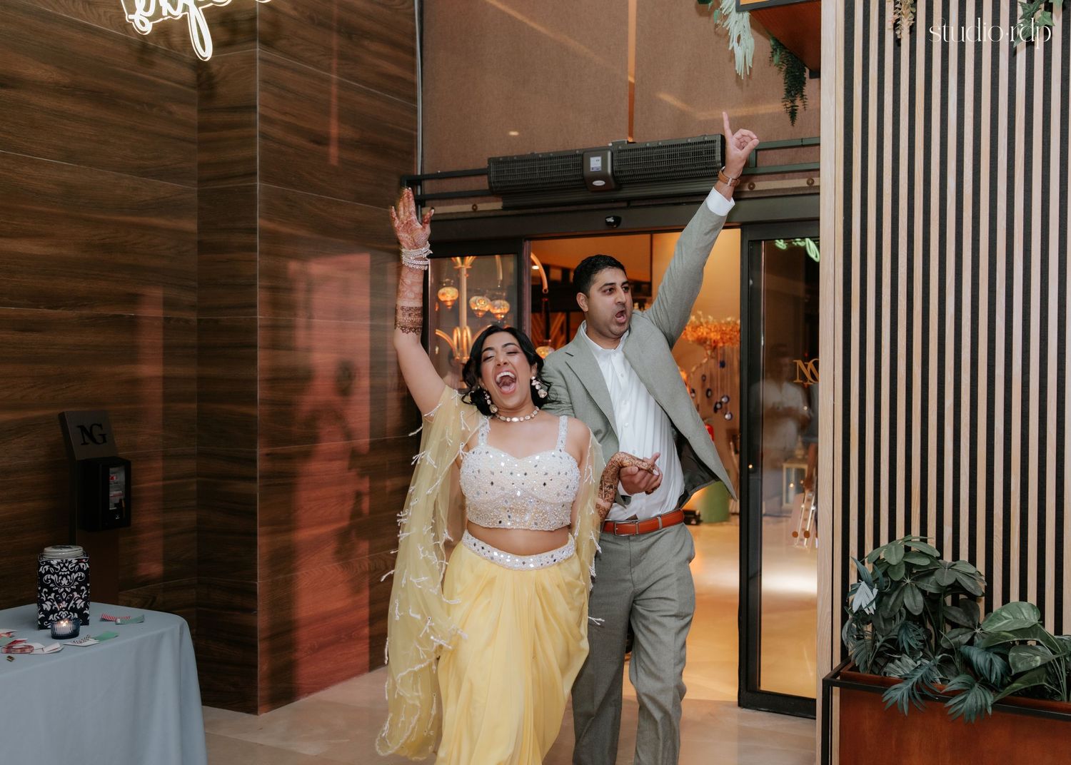 Bride in yellow lehenga and groom in gray suit celebrate grand entrance at wedding reception.