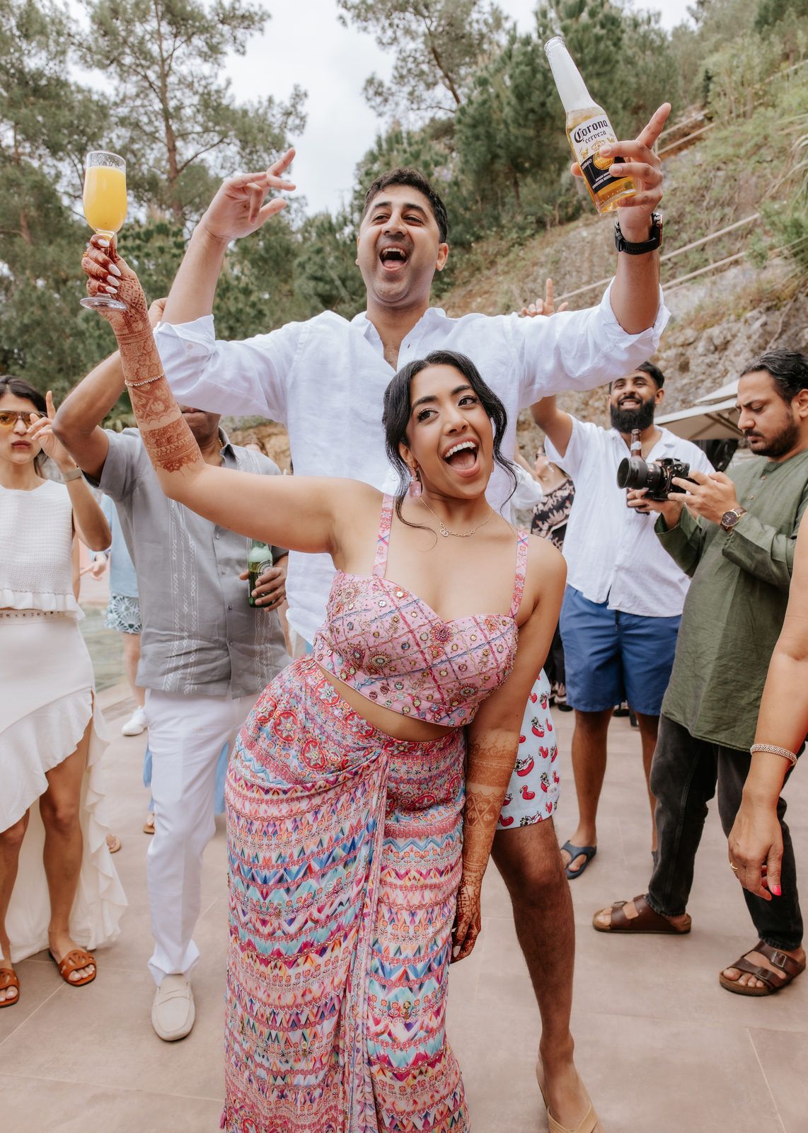 Couple celebrating at outdoor party, woman in colorful sari dancing joyfully while man holds Corona beer bottle overhead.