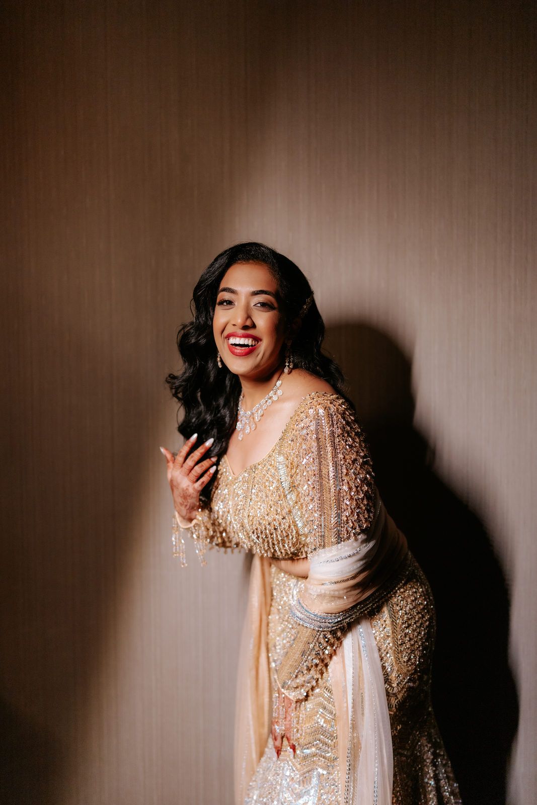 A woman in a gold embroidered lehenga smiles joyfully against a warm beige wall.