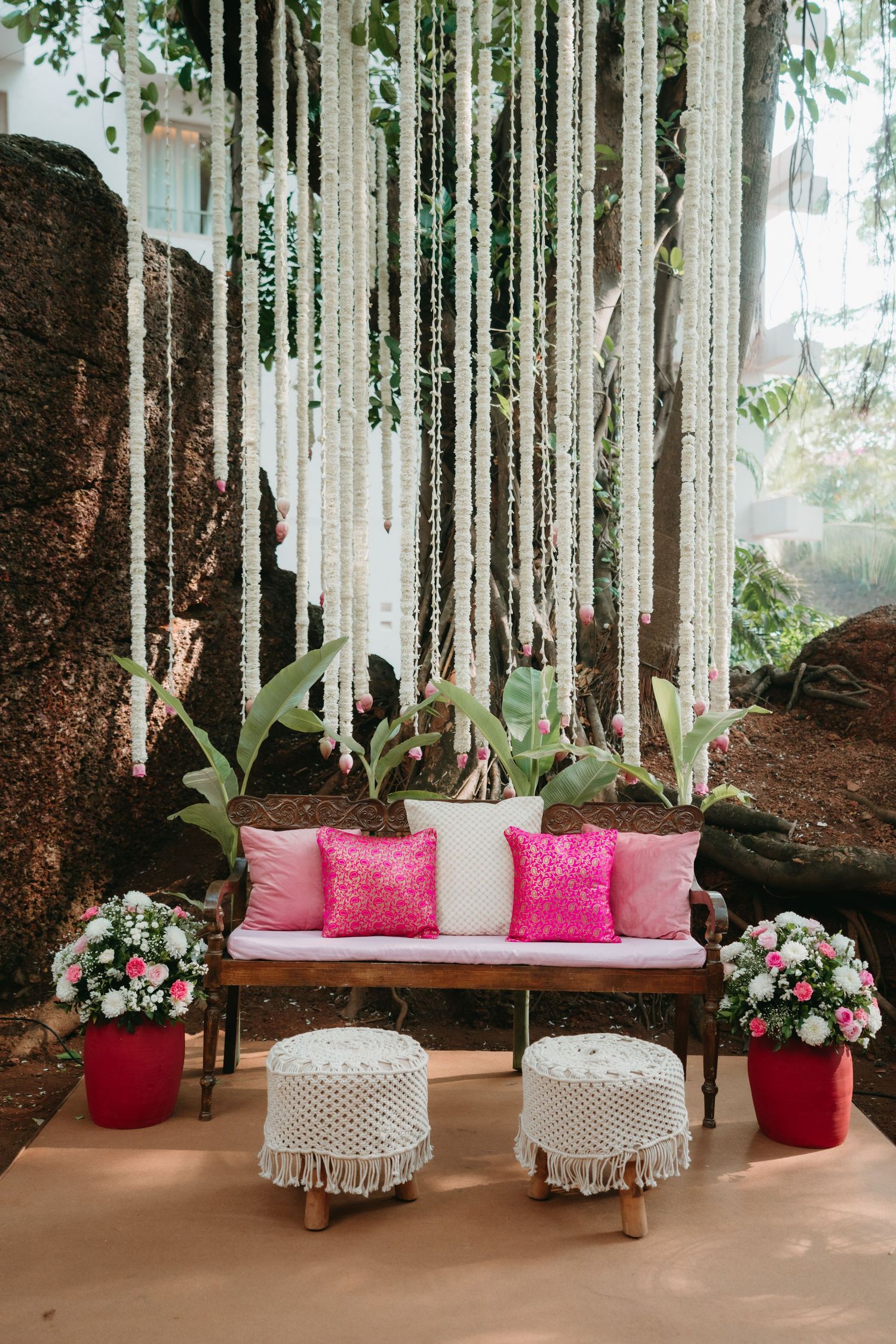 Boho wedding lounge with wooden bench, pink pillows, macrame ottomans, floral arrangements, and white hanging decorations outdoors.