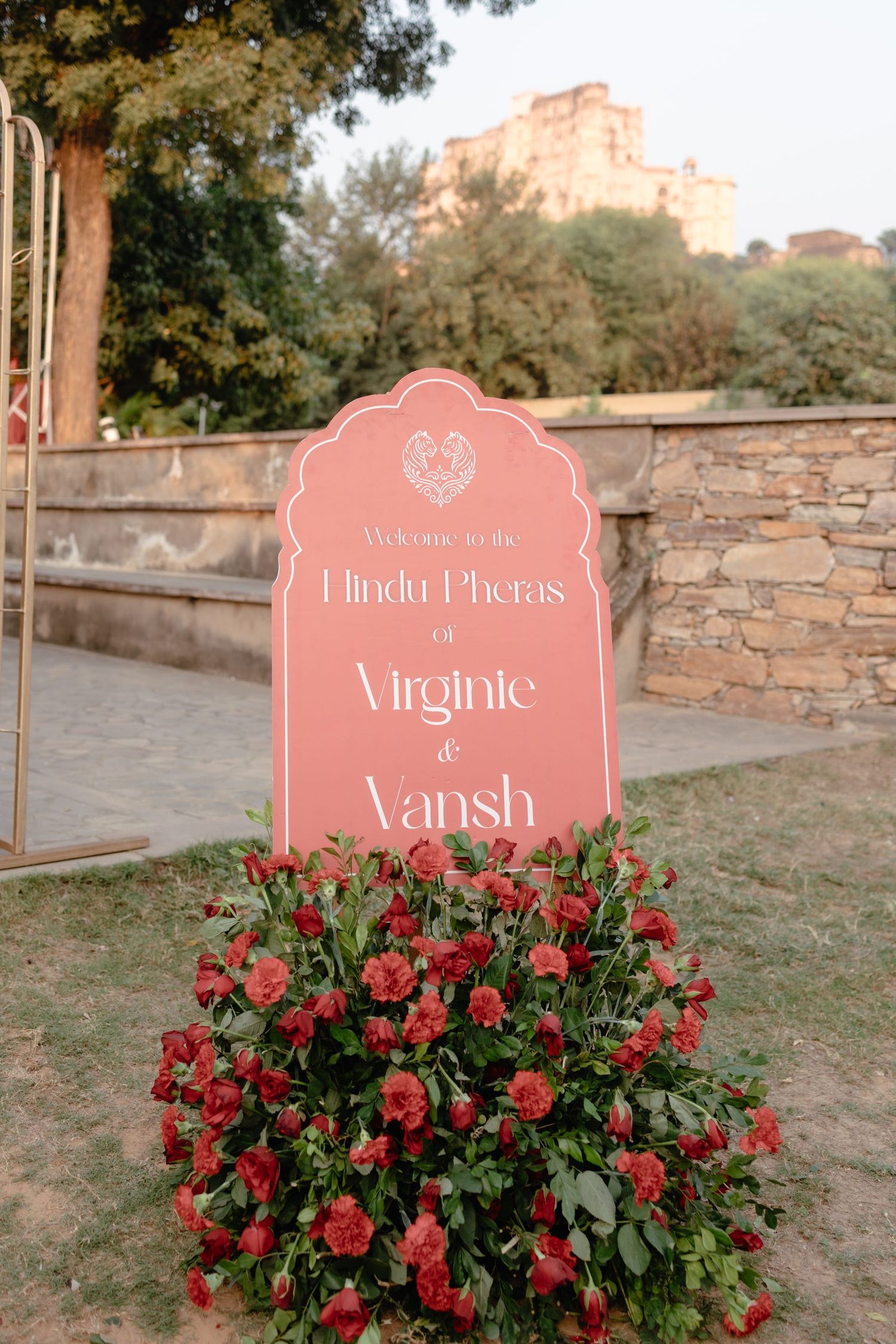 Pink wedding signage with floral decorations displaying names against stone wall.