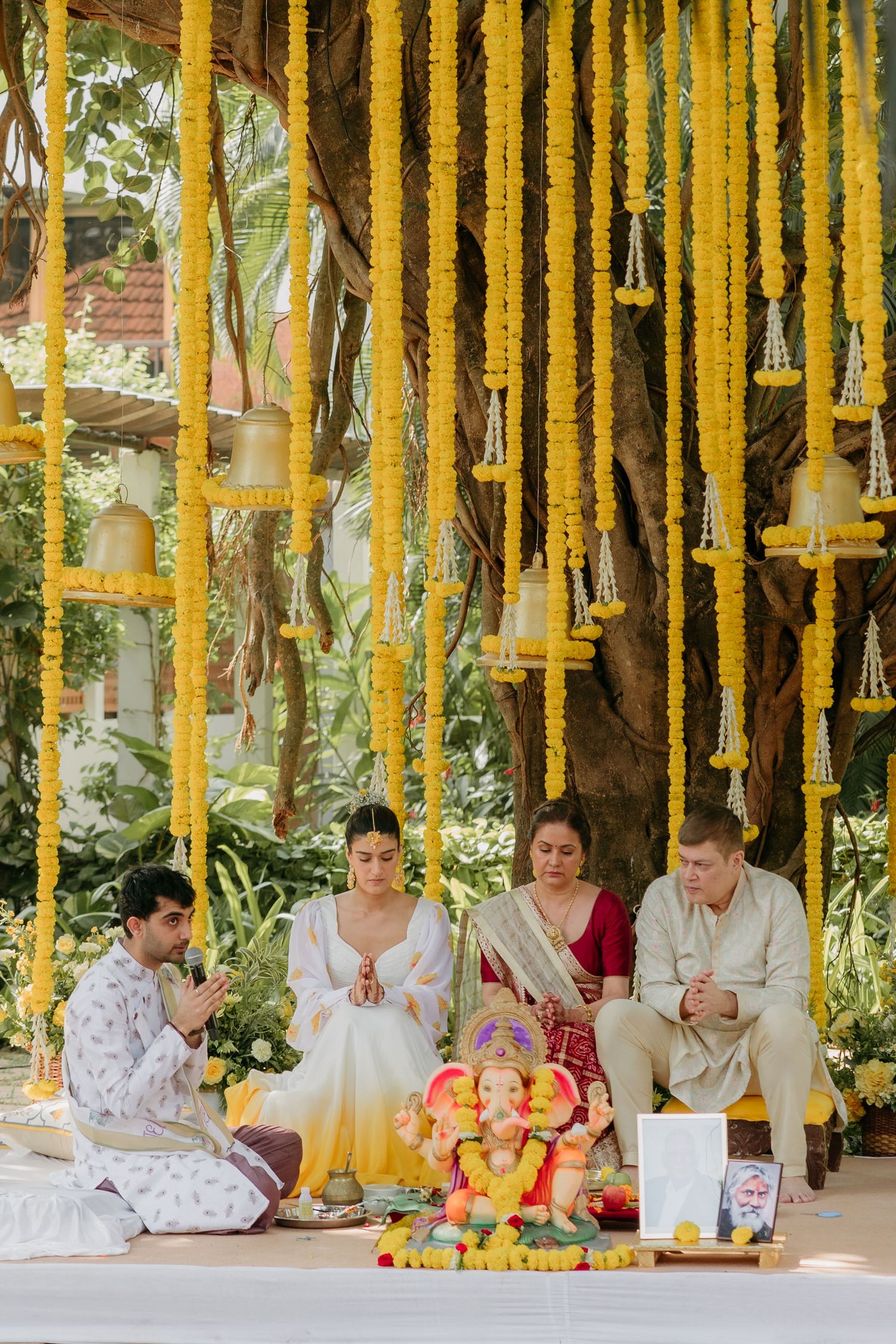 Indian wedding ceremony with Ganesh idol, yellow marigold garlands, and four people in traditional attire praying outdoors.