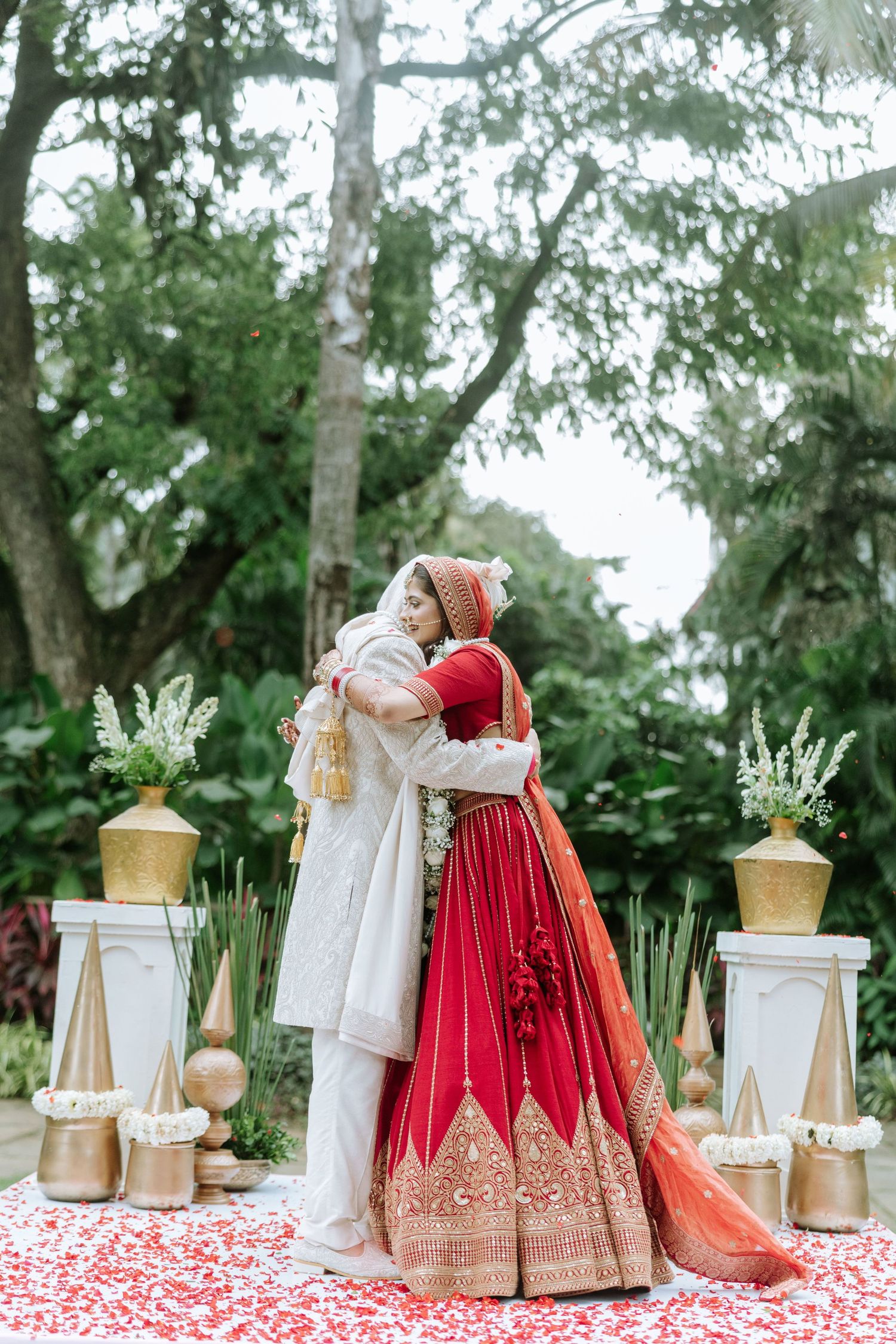 Indian bride in red lehenga and groom in white sherwani share a warm embrace during outdoor wedding ceremony.