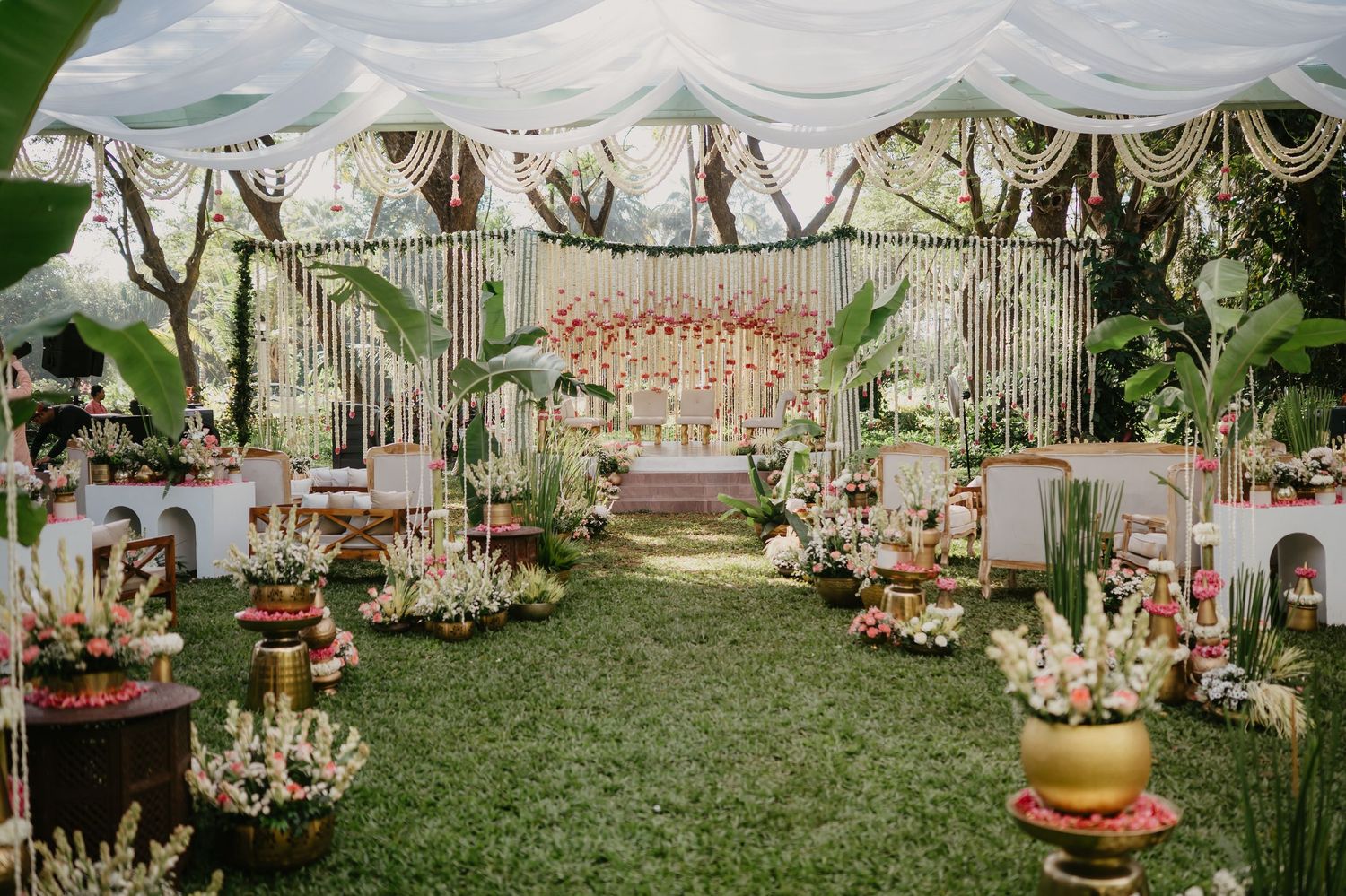 Outdoor wedding reception setup with draped white canopy, floral arrangements and greenery on a lush grass lawn.