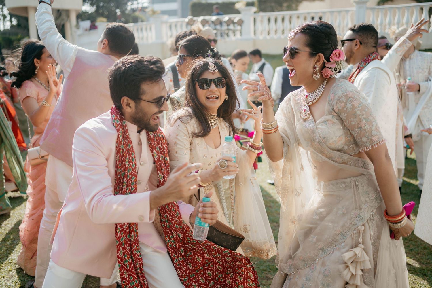 Guests in pink and red attire celebrate at an outdoor Indian daytime celebration with drinks and dancing.