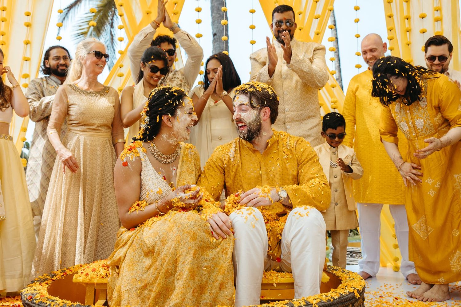 Joyful Indian wedding Haldi ceremony with couple in yellow outfits surrounded by celebrating guests throwing flowers.