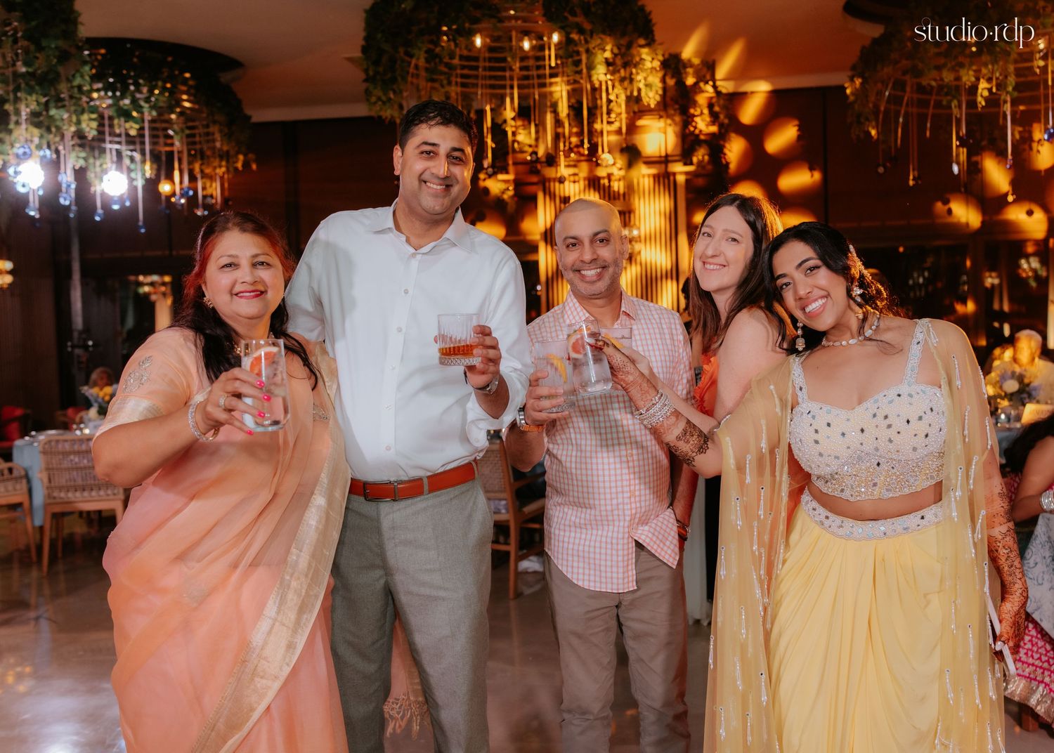 Group of five people smiling and raising glasses in a toast at a colorful Indian wedding celebration.