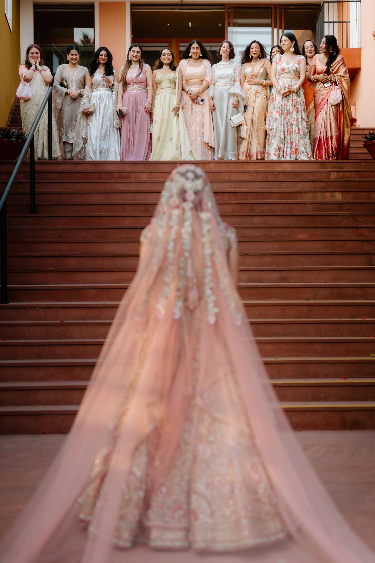 A group in pastel pink and white formal dresses poses elegantly on wooden stairs during a wedding celebration.