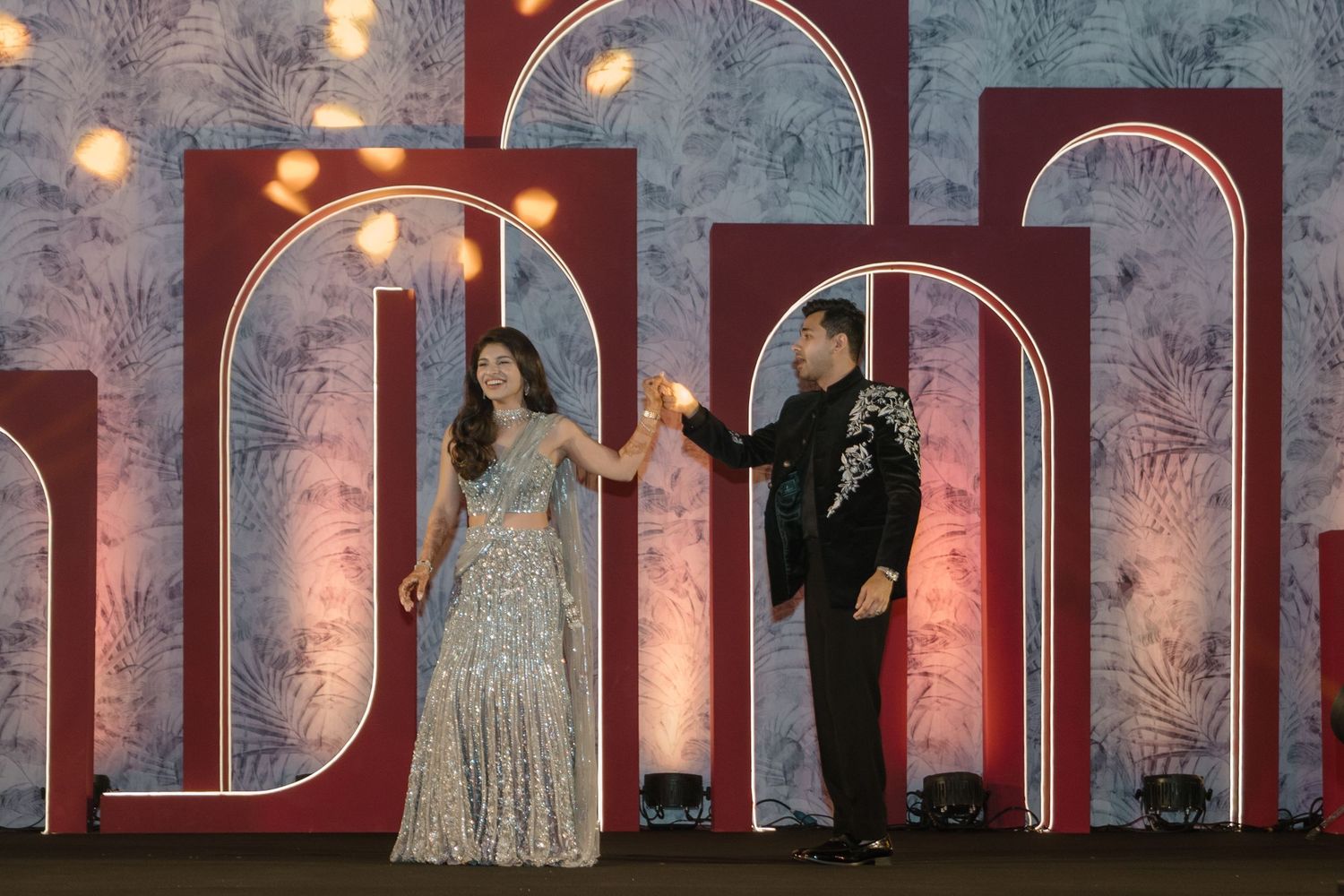 Elegant couple poses in front of decorative arches with glowing lights and floral patterns at formal event.
