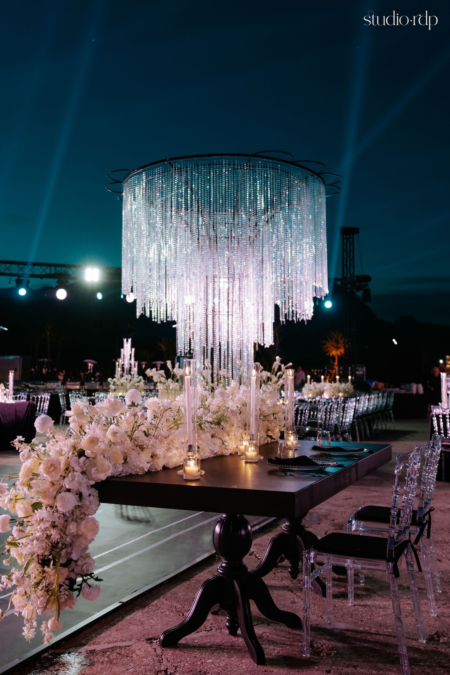 Elegant outdoor wedding reception with crystal chandelier, white floral arrangements, and ghost chairs at night.