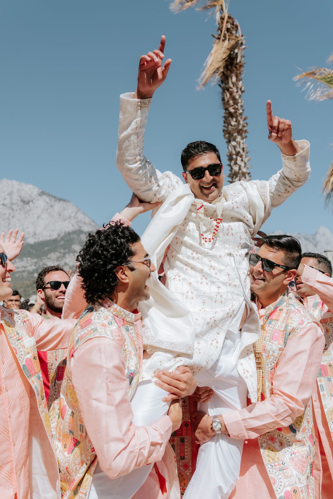 Groom in white sherwani lifted by groomsmen in pink kurtas during a joyful baraat celebration.