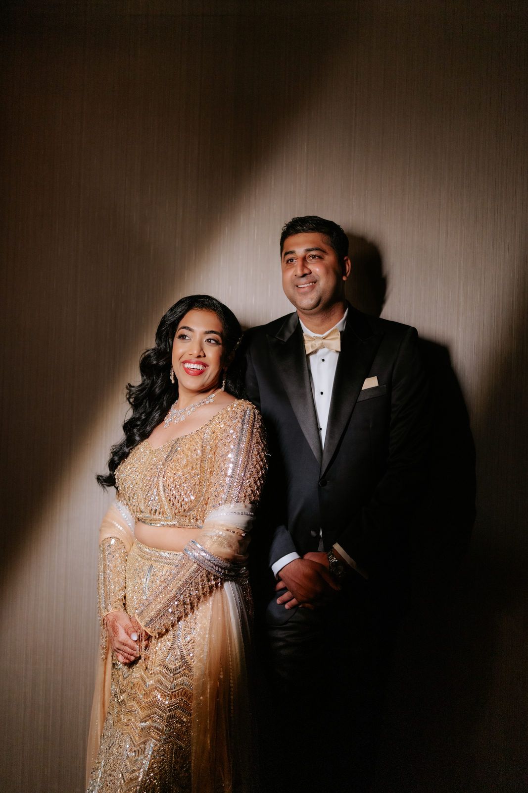 Elegant couple posing together, bride in gold embroidered lehenga and groom in black tuxedo at Indian wedding reception.