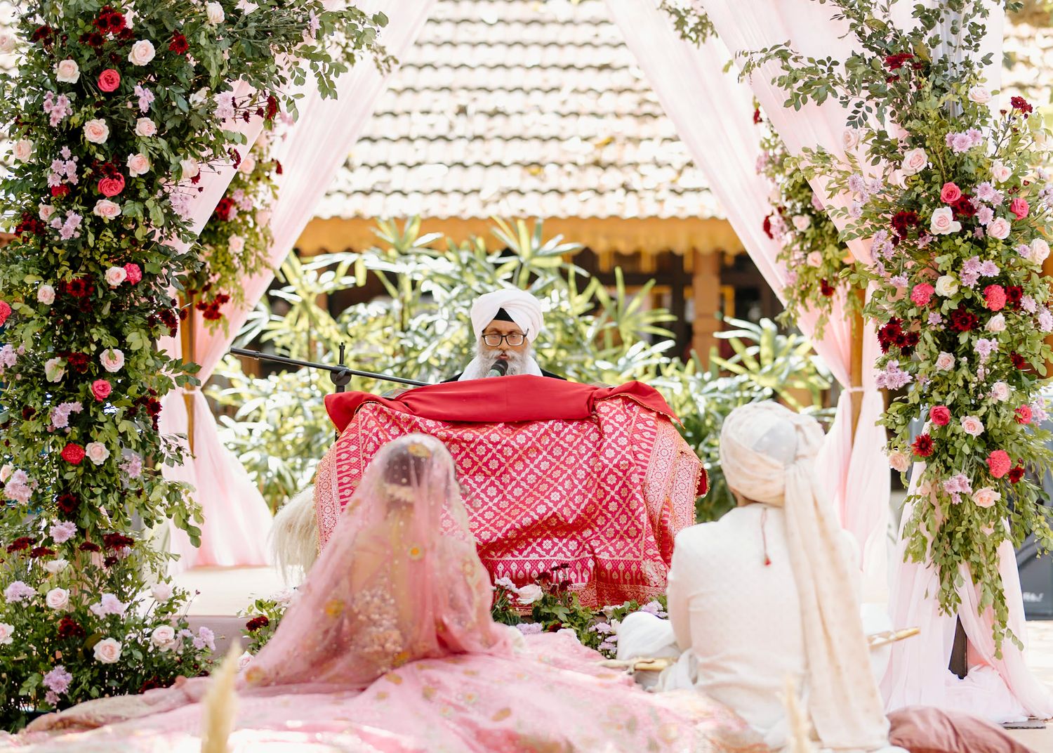 Couple seated at floral mandap decorated with pink and red roses during traditional Indian wedding ceremony.